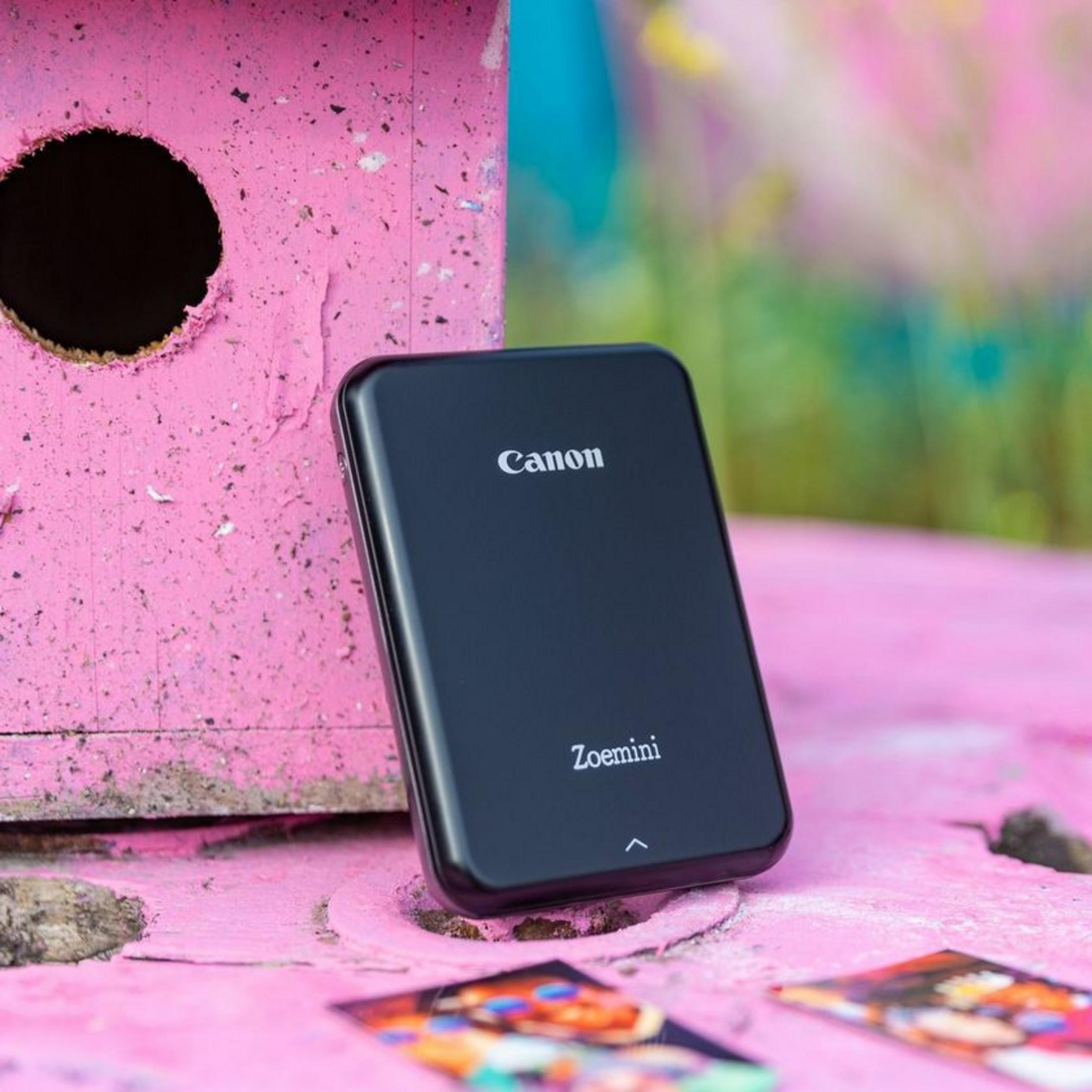 Una impresora portátil Canon Zoemini negra sobre una superficie rosa con una casita de pájaros rosa al fondo.