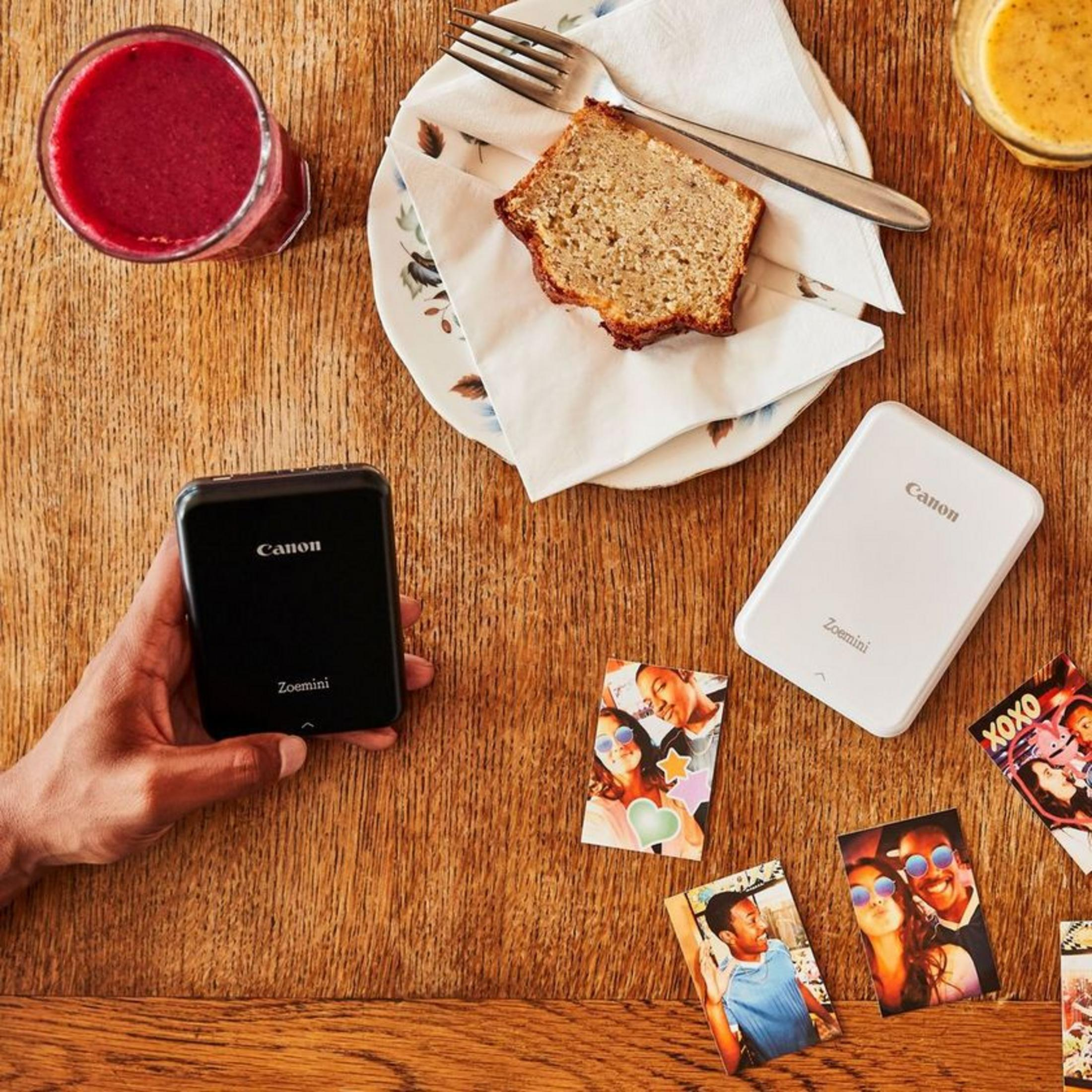 Ręce trzymające dwie drukarki Canon Zoemini i zdjęcia na drewnianym stole. Napoje i jedzenie obecne.