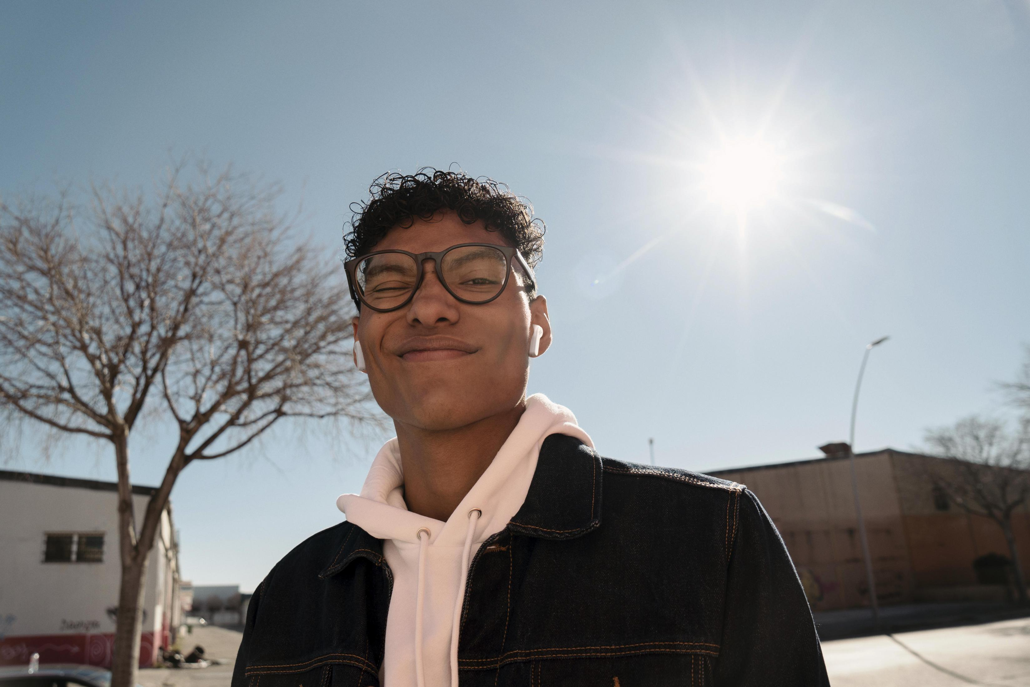 Uśmiechnięty mężczyzna w okularach i słuchawkach, stojący na zewnątrz w słoneczny dzień.