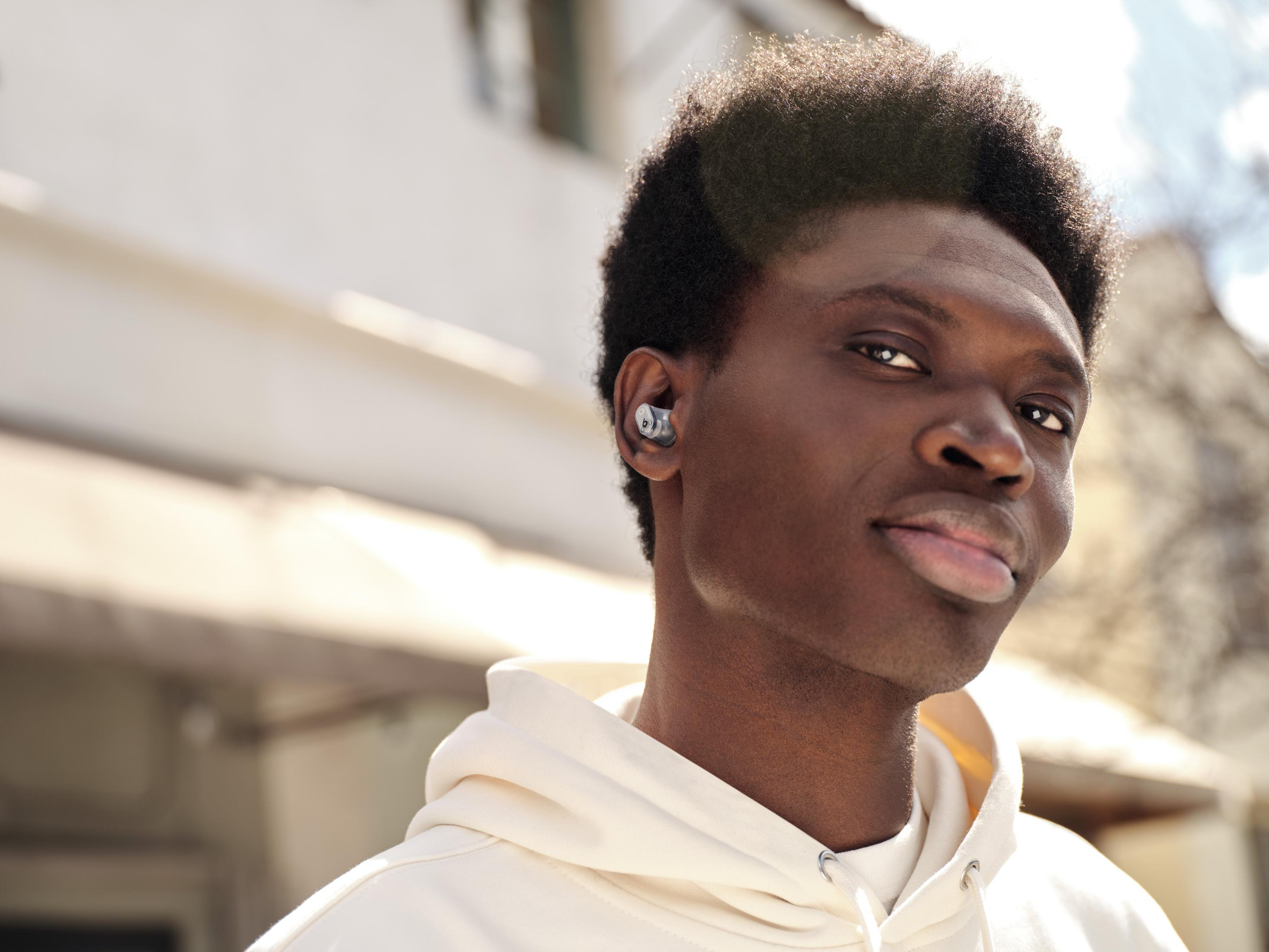 Czarny mężczyzna w białej bluzie z kapturem, ze słuchawką w uchu. Uśmiechnięty.