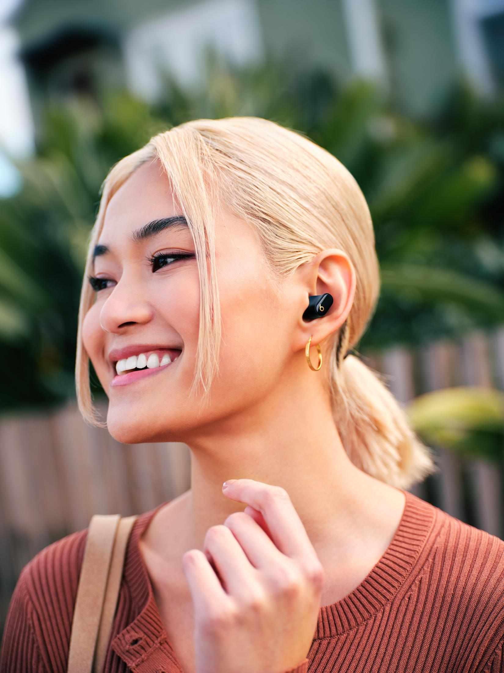 Kobieta z blond włosami uśmiecha się, ma na sobie brązową koszulę i ma słuchawki. Zielone liście w tle.