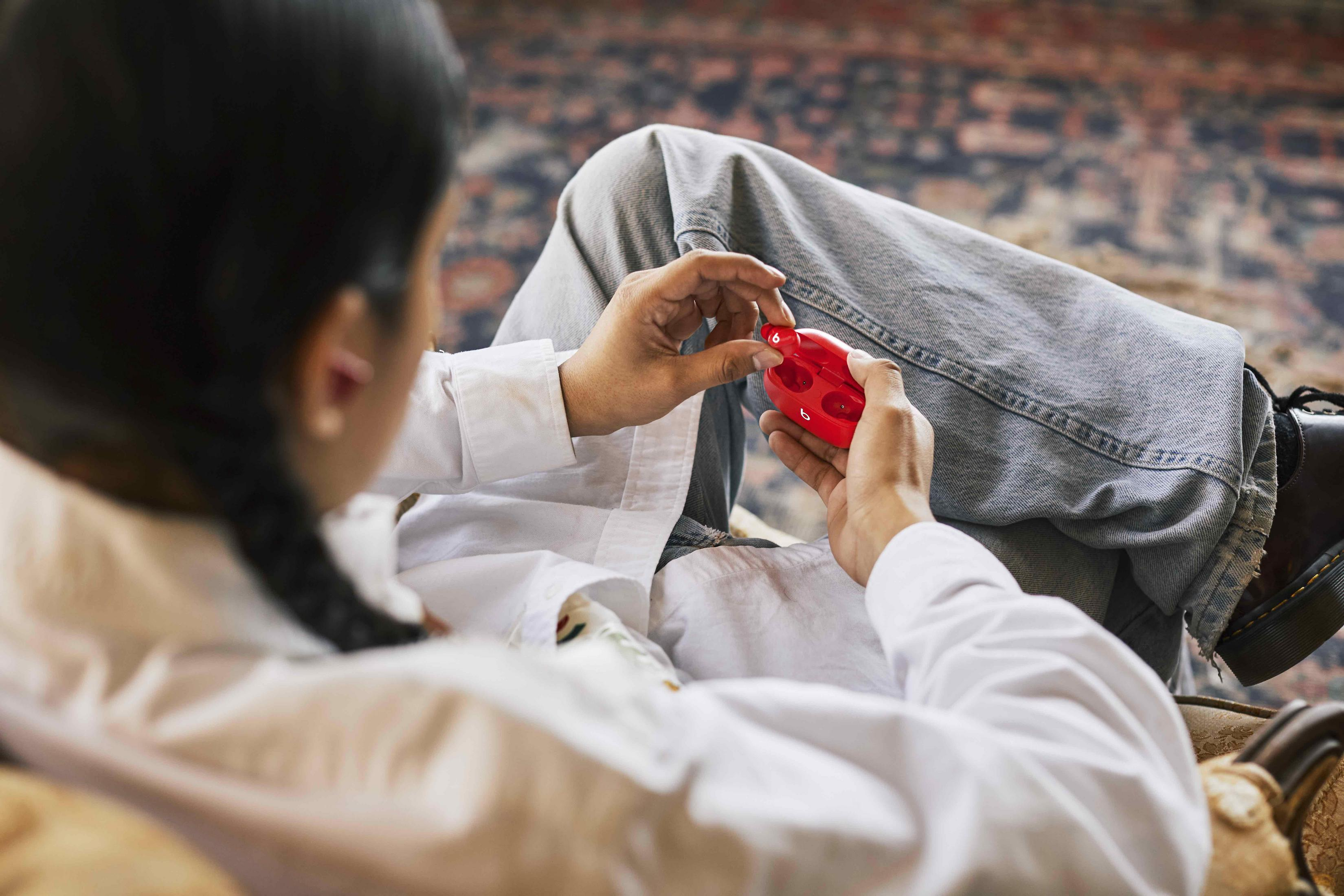 Osoba trzyma czerwone urządzenie, siedząc na kanapie z wzorzystym dywanem.