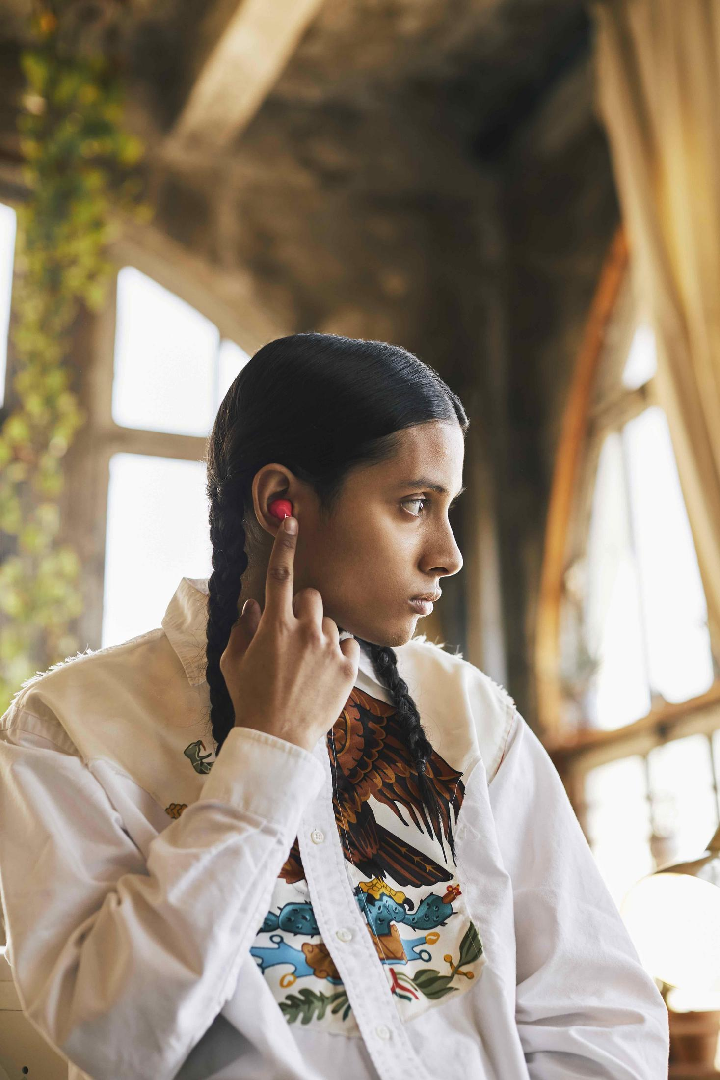 Osoba z ciemnymi włosami i warkoczami nosi słuchawki douszne, dotykając jednej z nich. Tło ma okno i rośliny.