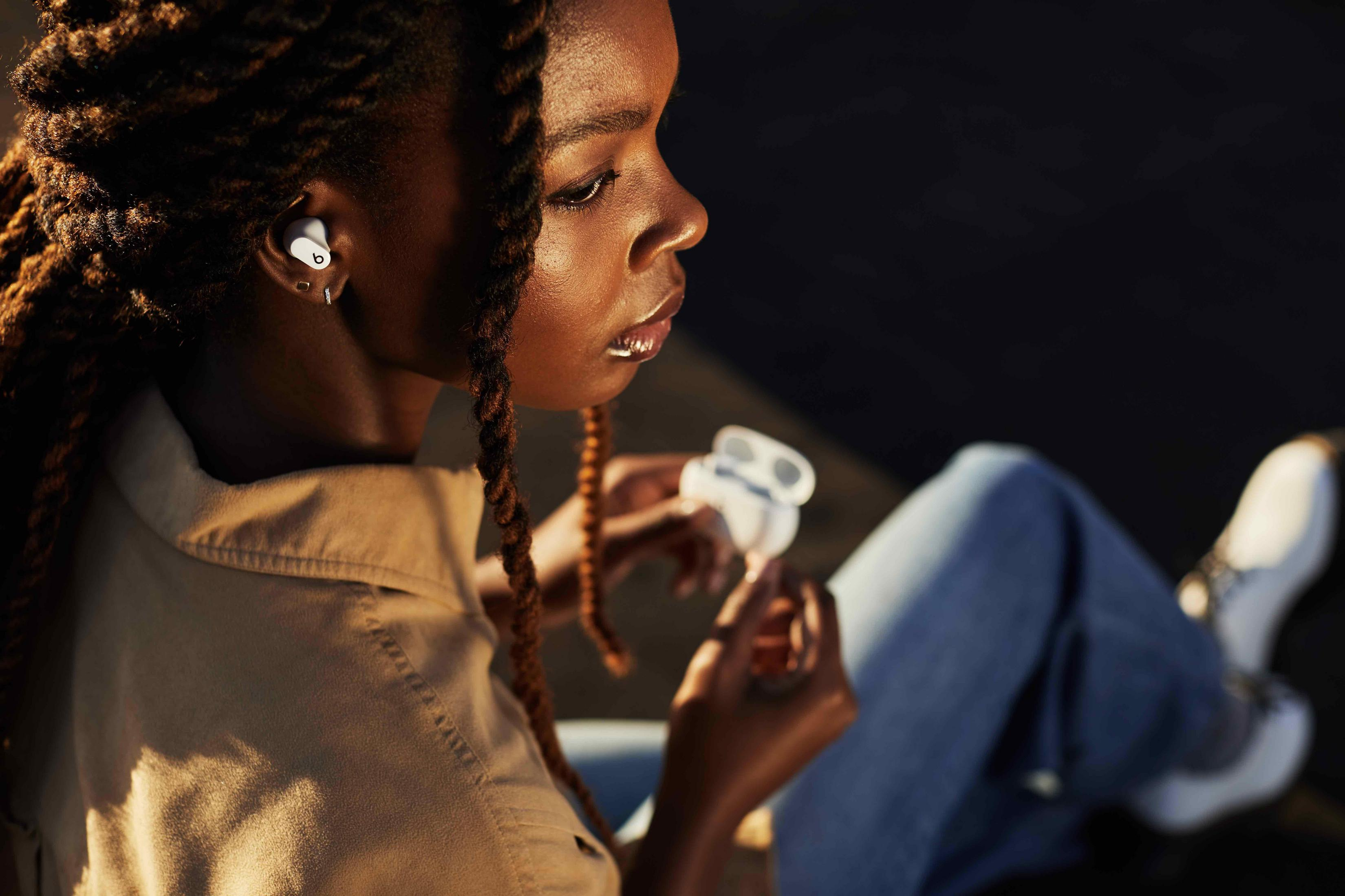 Kobieta w białych słuchawkach, trzymająca białe etui ładujące, siedząca w słońcu.