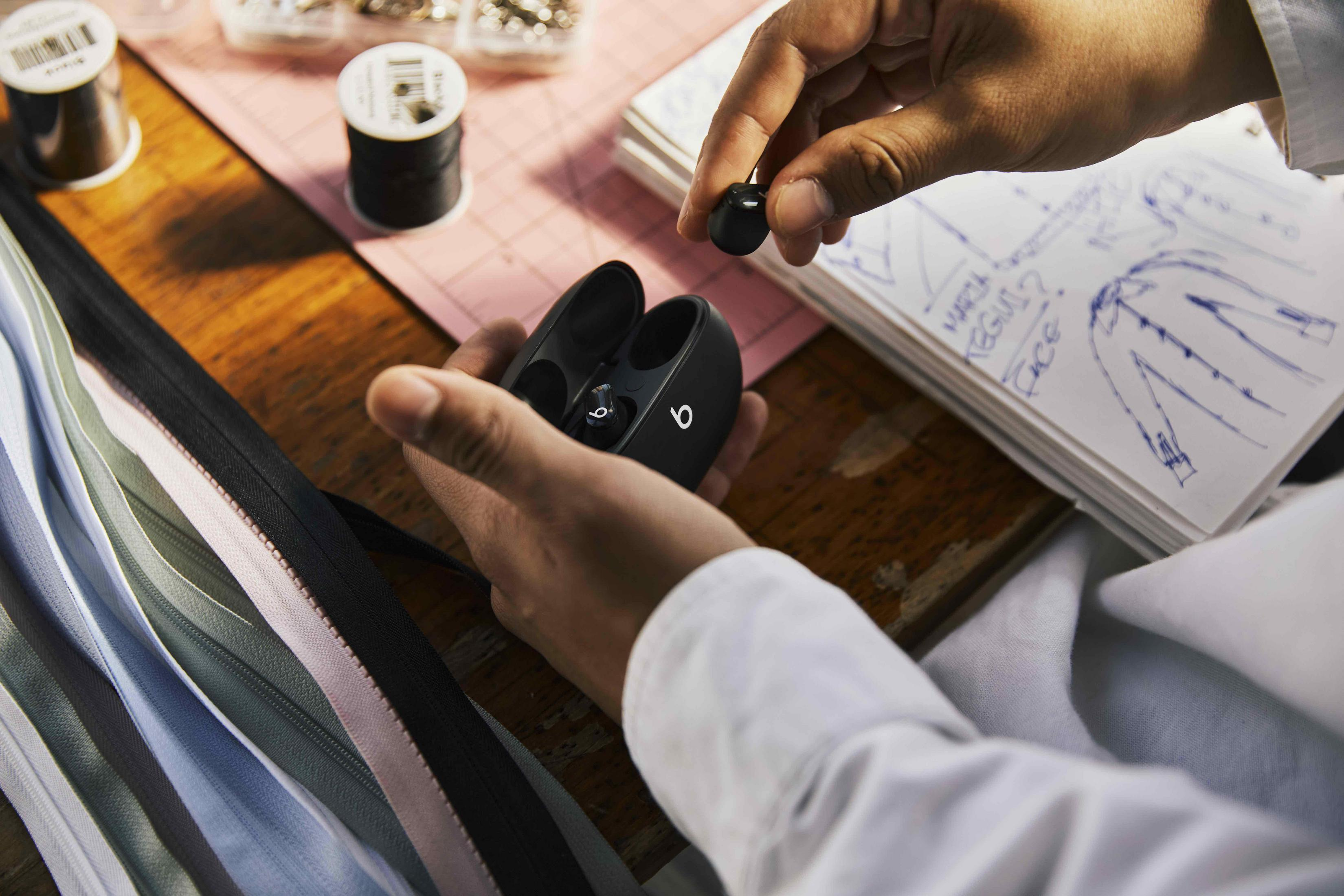 Ręce trzymające czarne słuchawki Beats obok czarnego etui. Tło: materiały do szycia i rysunki.