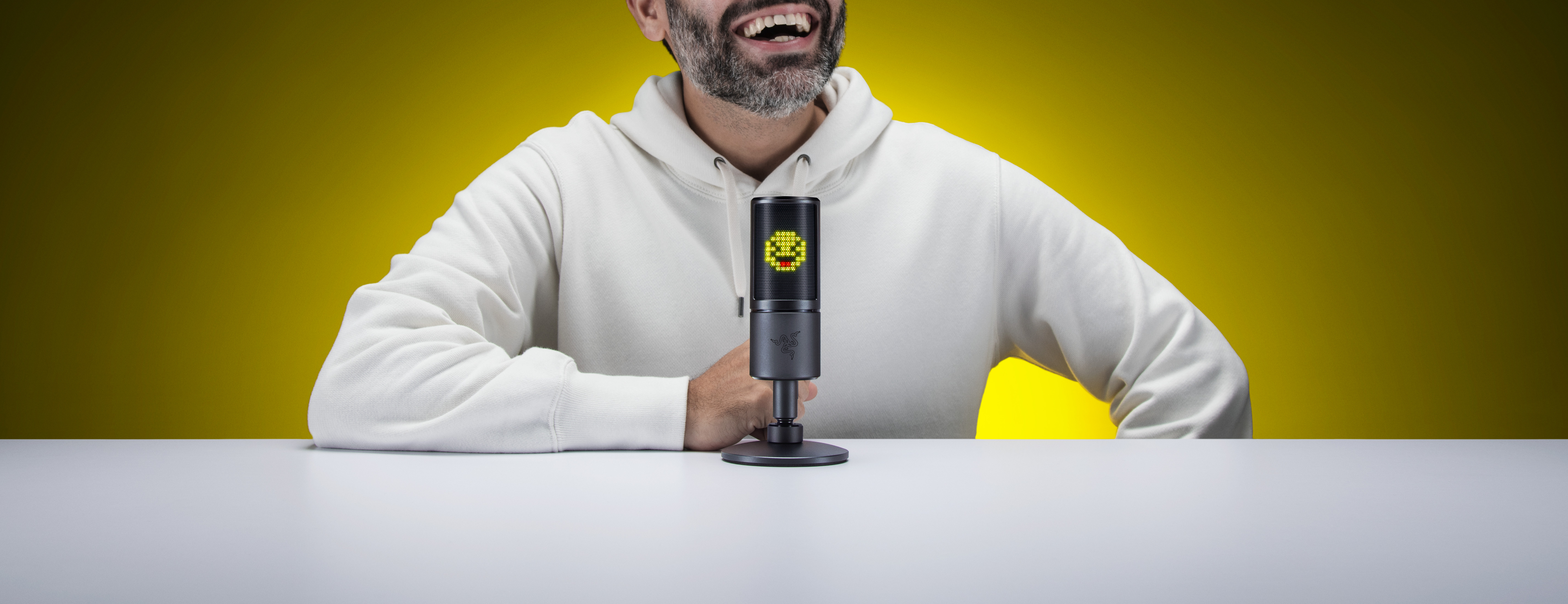 Mężczyzna z brodą uśmiechnięty, mikrofon na stole. Żółte tło. Ustawienia studia.