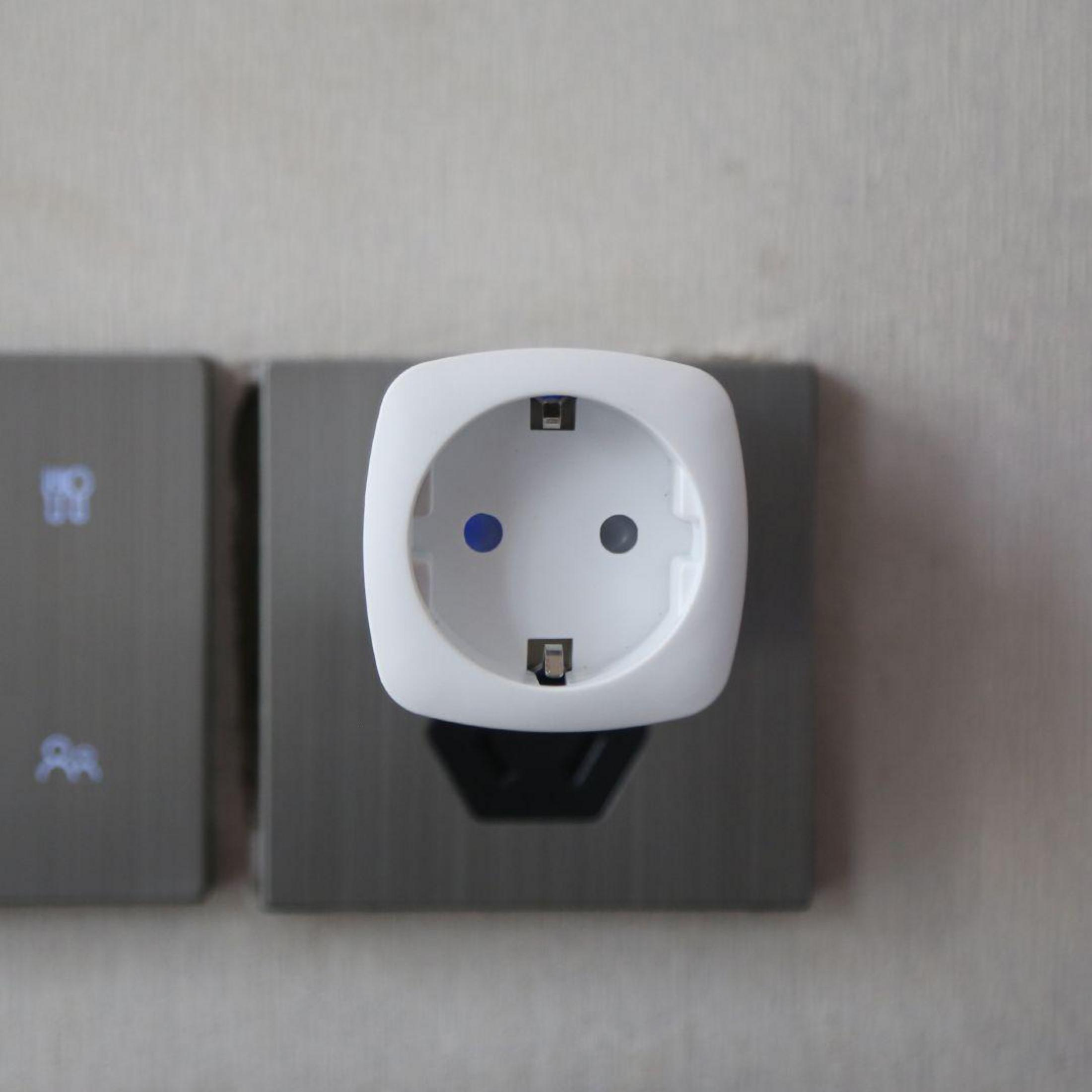 Een witte slimme stekker in een grijs stopcontact op een lichtgrijze muur.