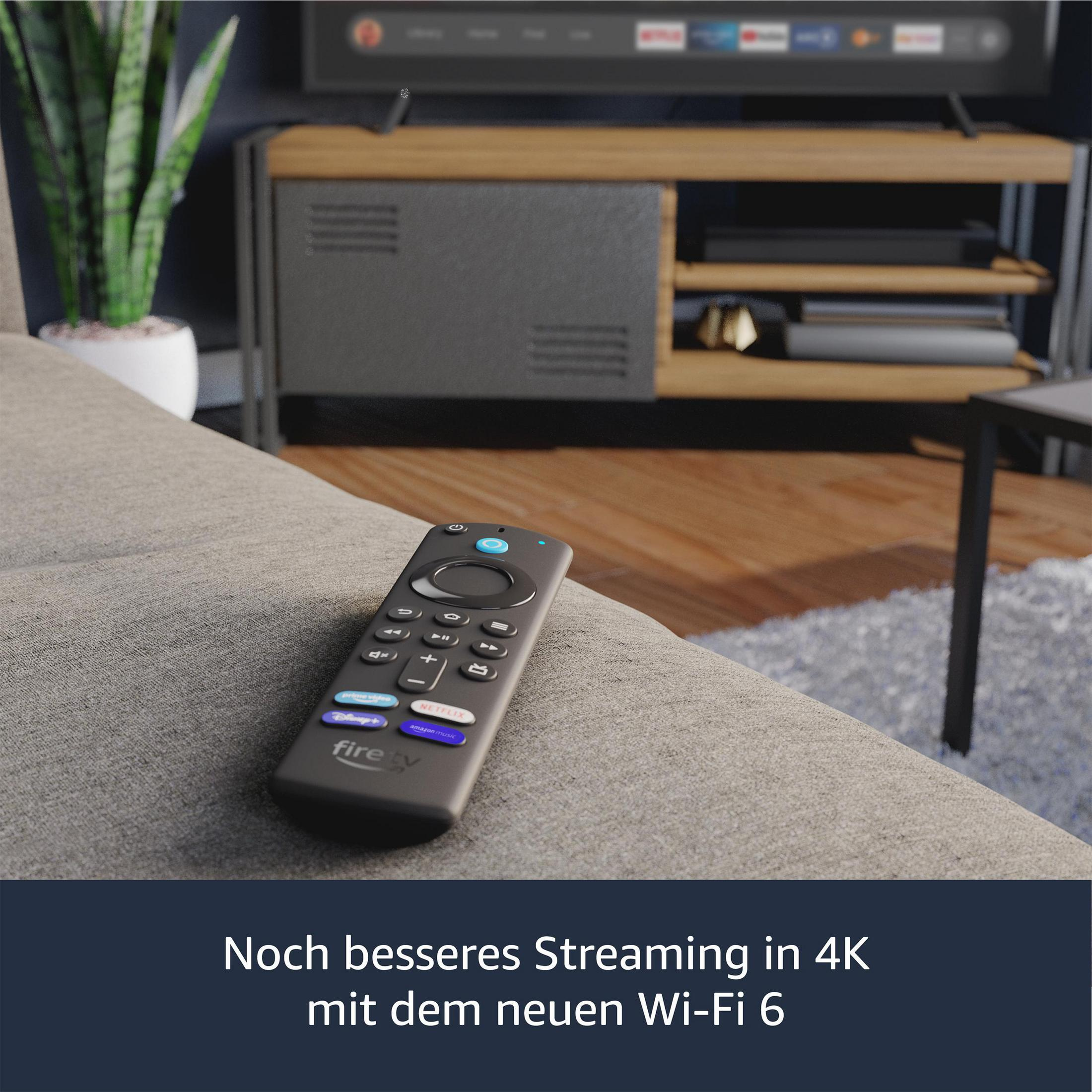 Eine schwarze Fire TV-Fernbedienung auf einem Sofa. Im Hintergrund ein Fernseher und ein Schrank.