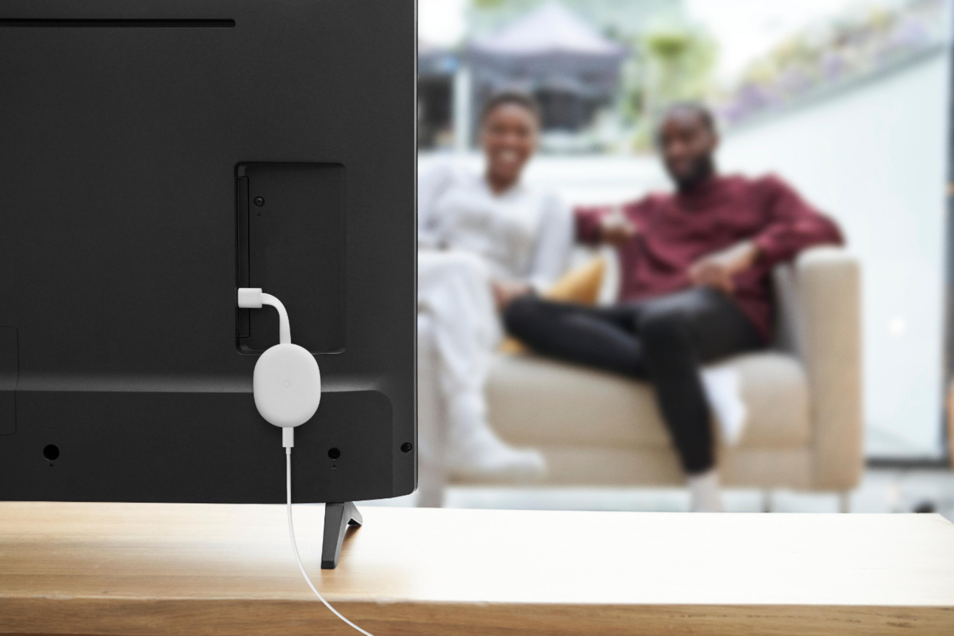 Ein Fernseher mit angeschlossenem Chromecast. Zwei Personen sitzen auf einem Sofa, unscharf im Hintergrund.