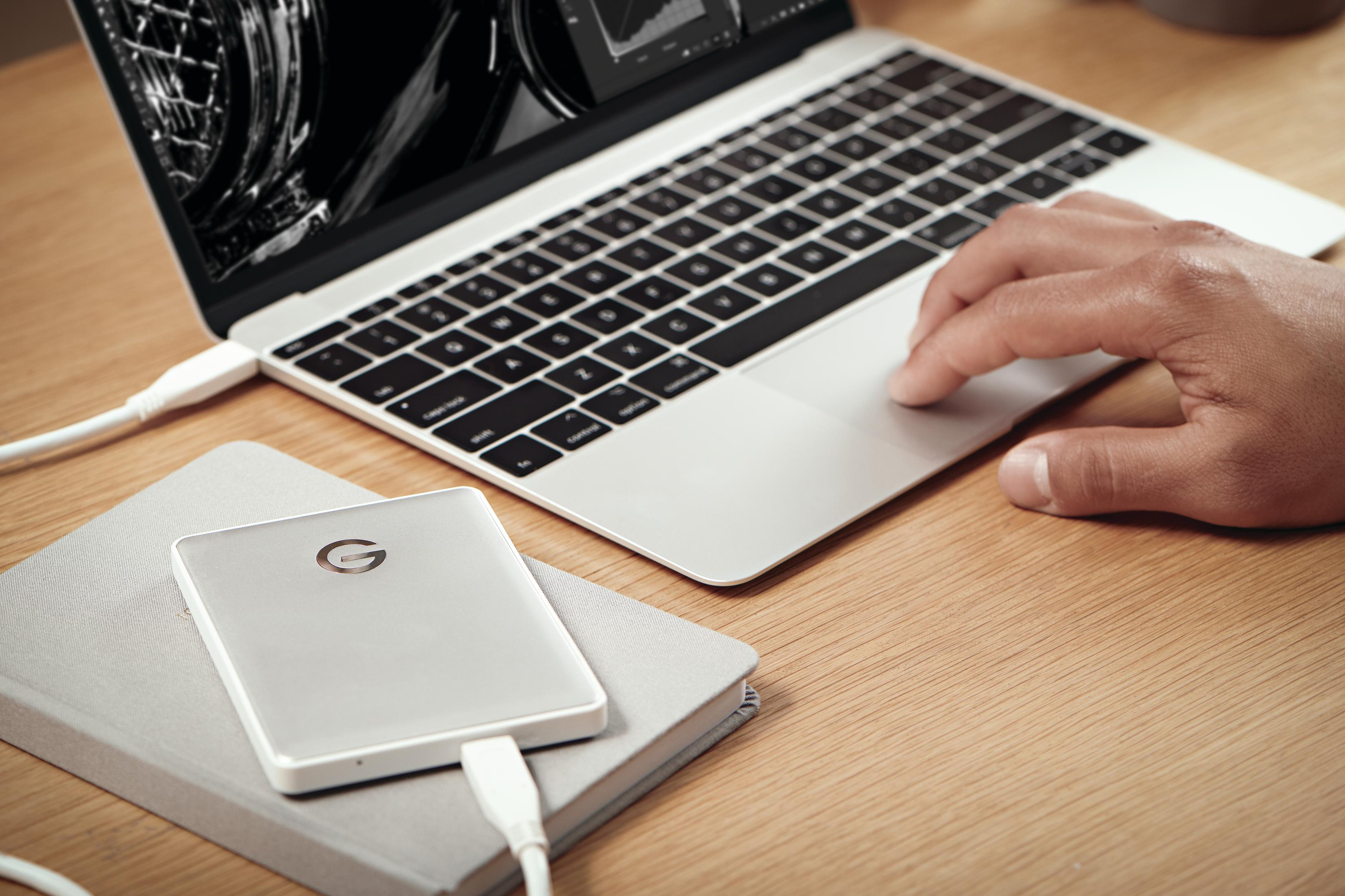 Un laptop con una mano sul trackpad, un disco rigido esterno argentato e un taccuino grigio.