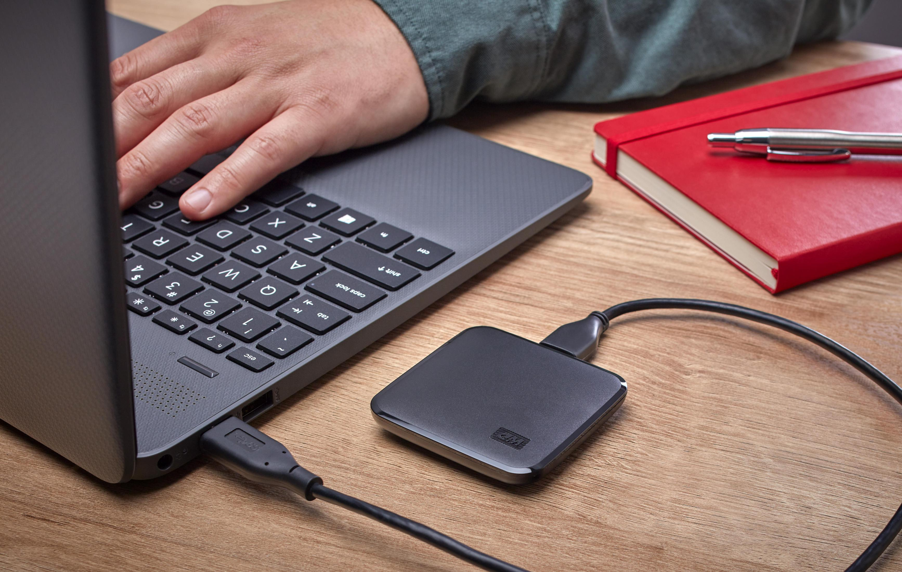 Osoba używa laptopa, podłączony dysk twardy. Czerwony notatnik i długopis po prawej.