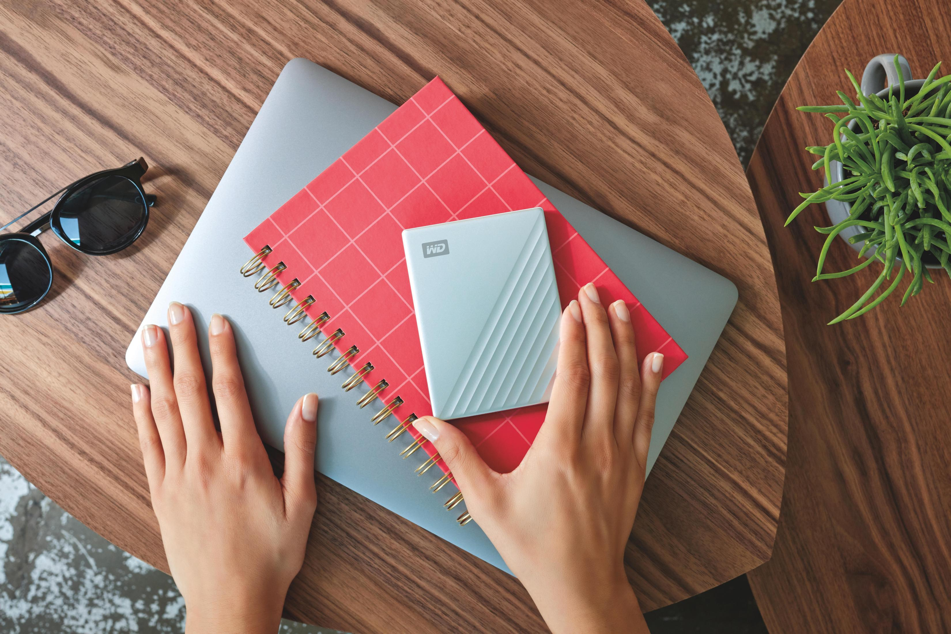 Ręce na laptopie i czerwonym notatniku, z zewnętrznym dyskiem twardym na górze, długopisem i rośliną.