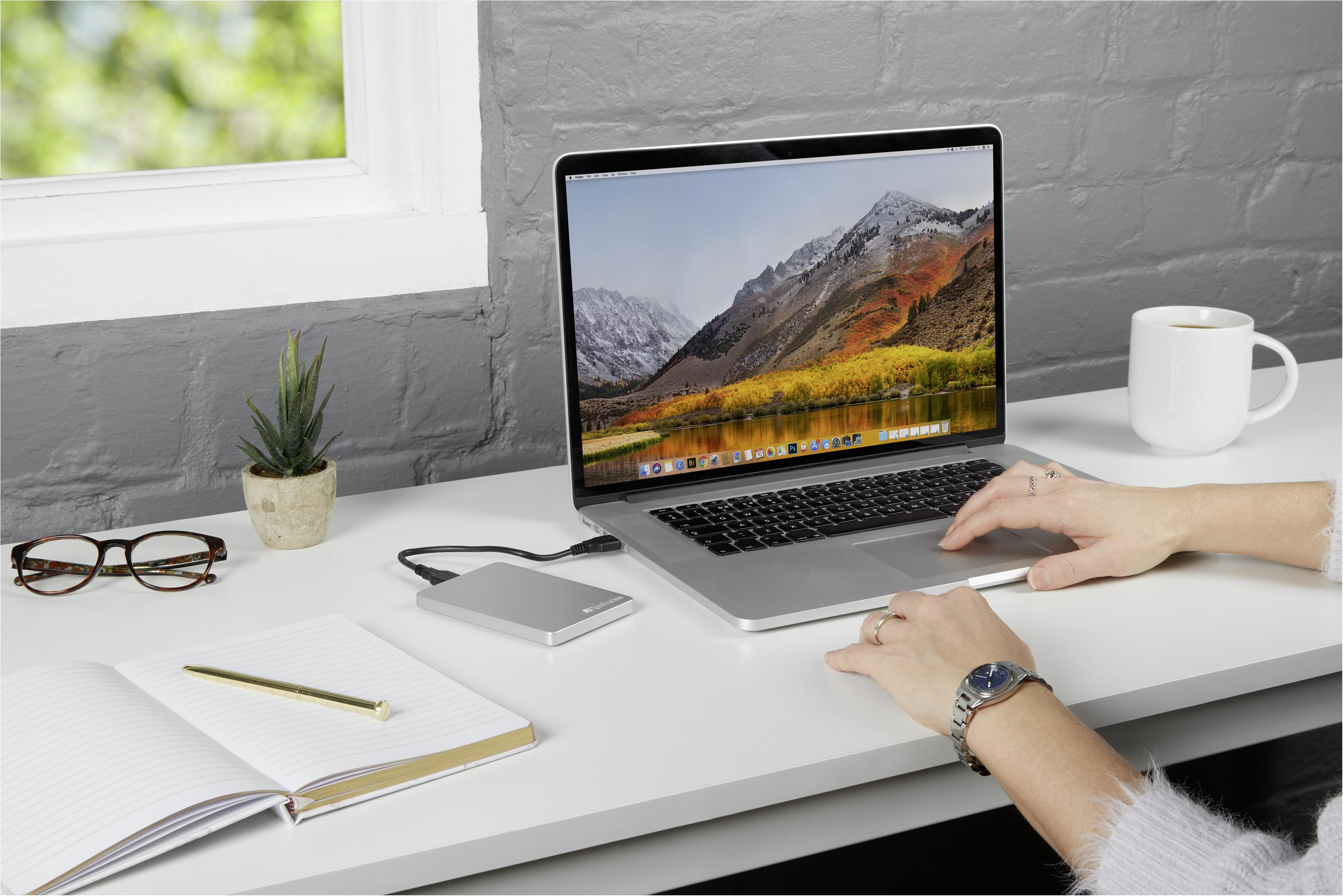 Laptop na białym biurku z podłączonym srebrnym zewnętrznym dyskiem twardym. Kobieta pisze.