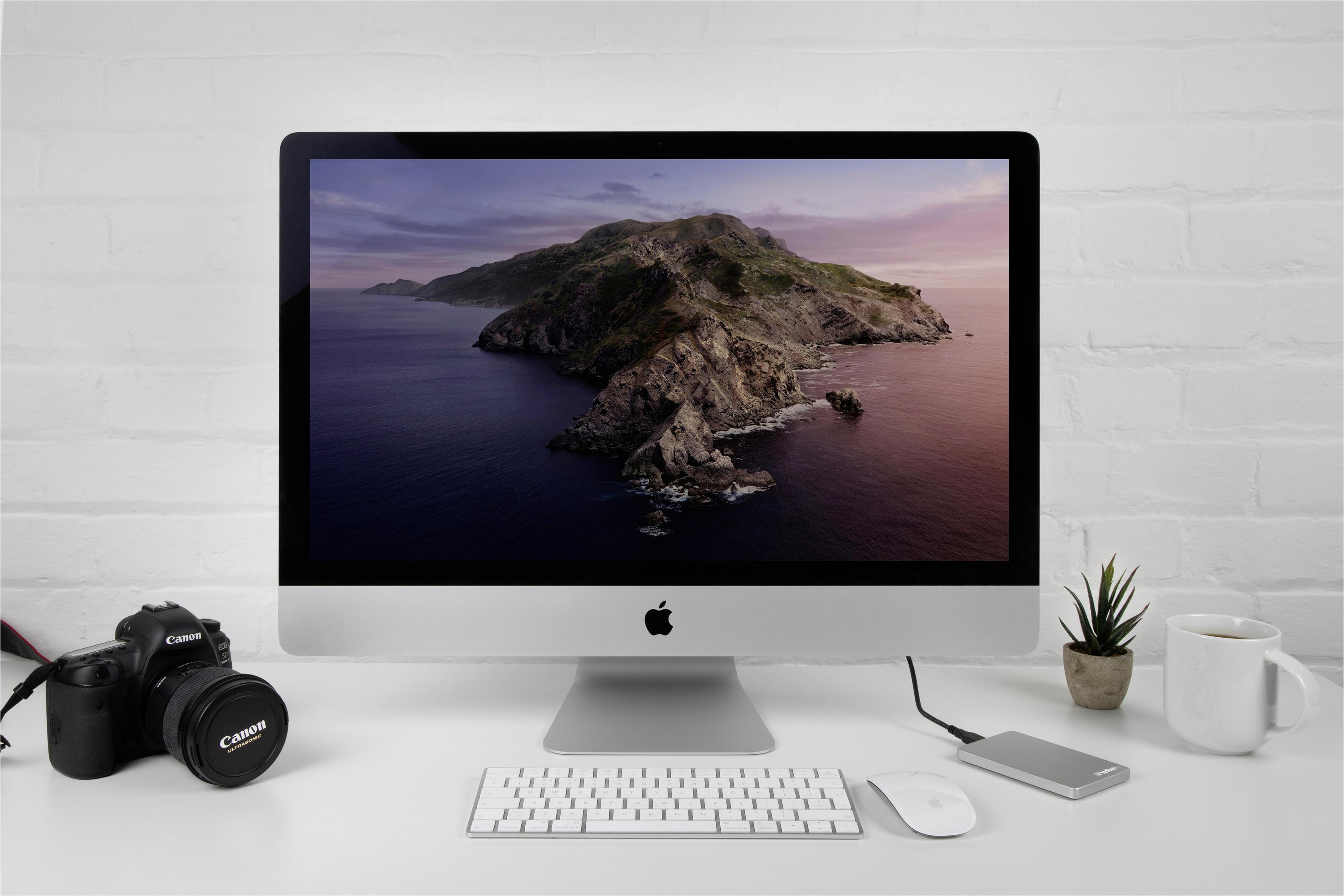 Komputer iMac z ekranem pokazującym krajobraz nadmorski, z aparatem, klawiaturą i kubkiem.