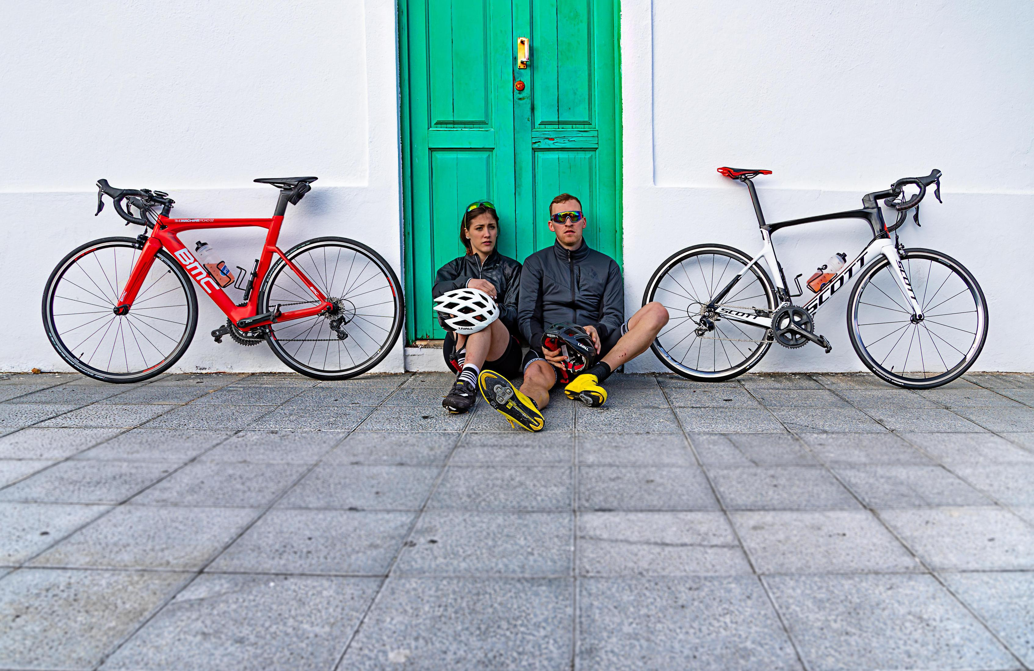Zwei Radfahrer mit Rädern, eins rot, eins schwarz-weiß, vor einer Tür.