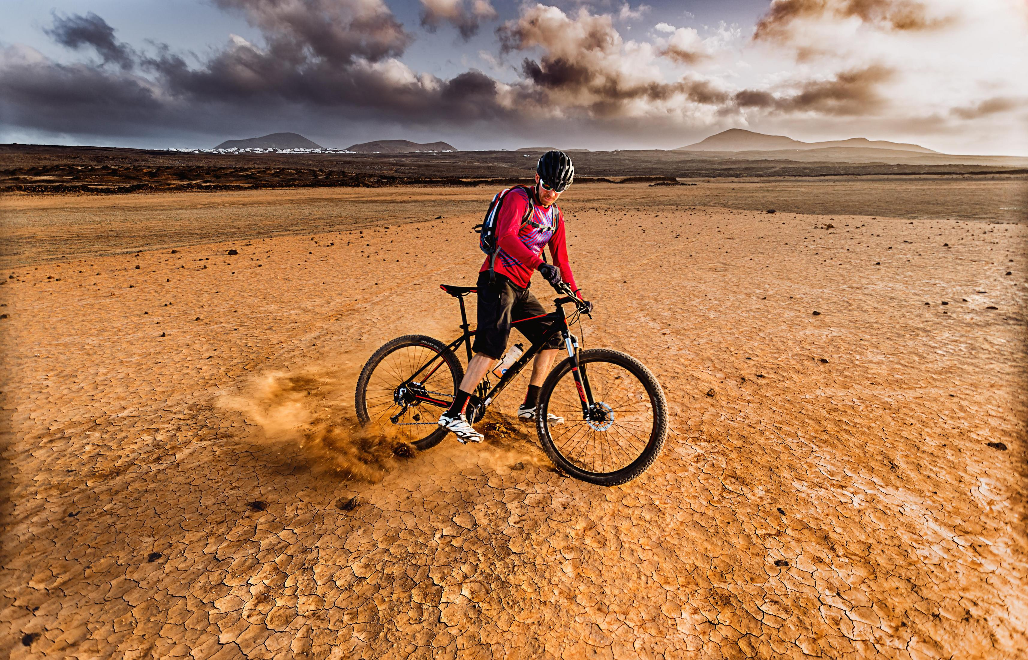 Radfahrer auf einem Mountainbike, der in einer Wüstenlandschaft Staub aufwirbelt. Der Himmel ist bewölkt.