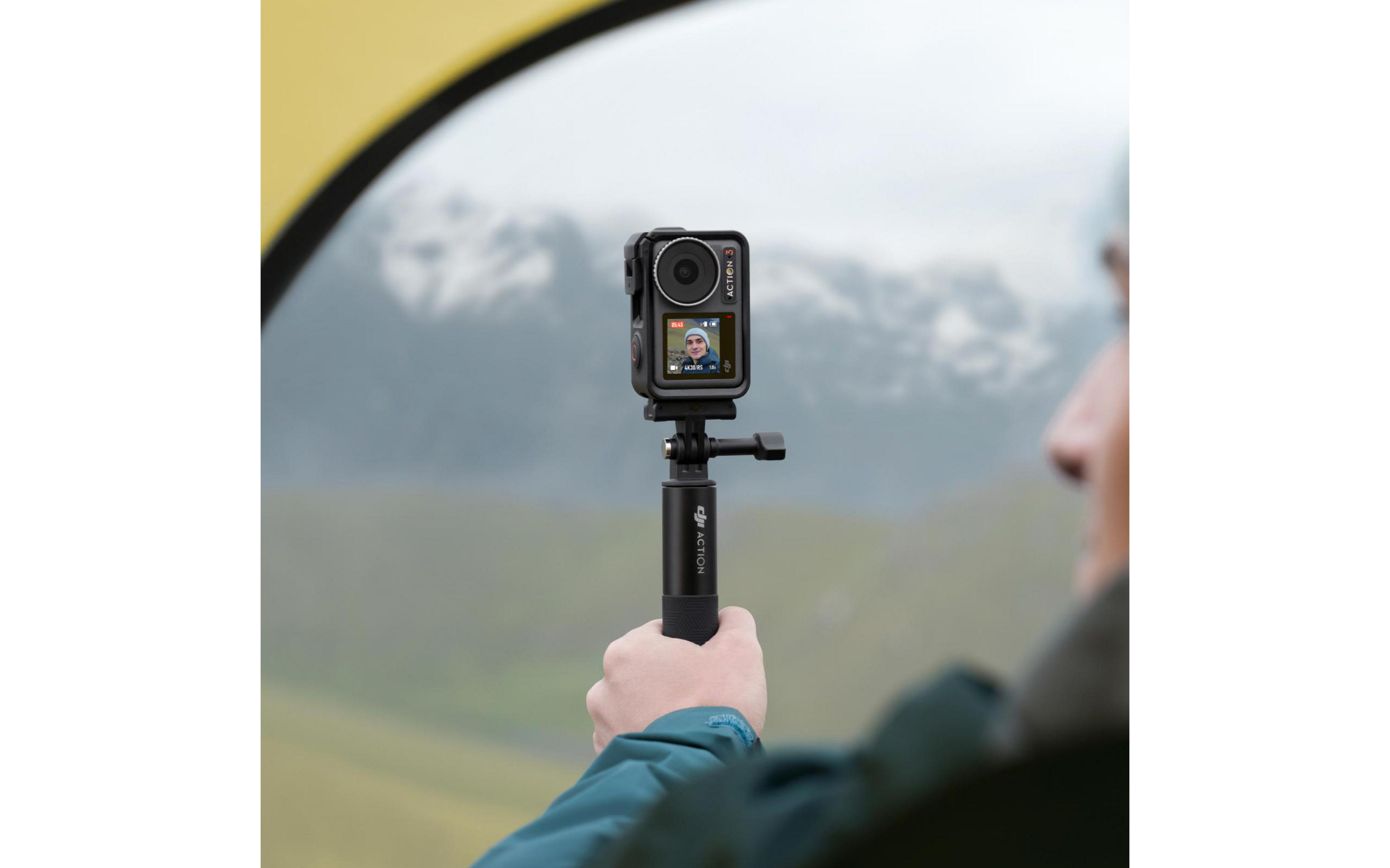 Osoba trzymająca aparat na patyku do selfie, z tłem krajobrazu.