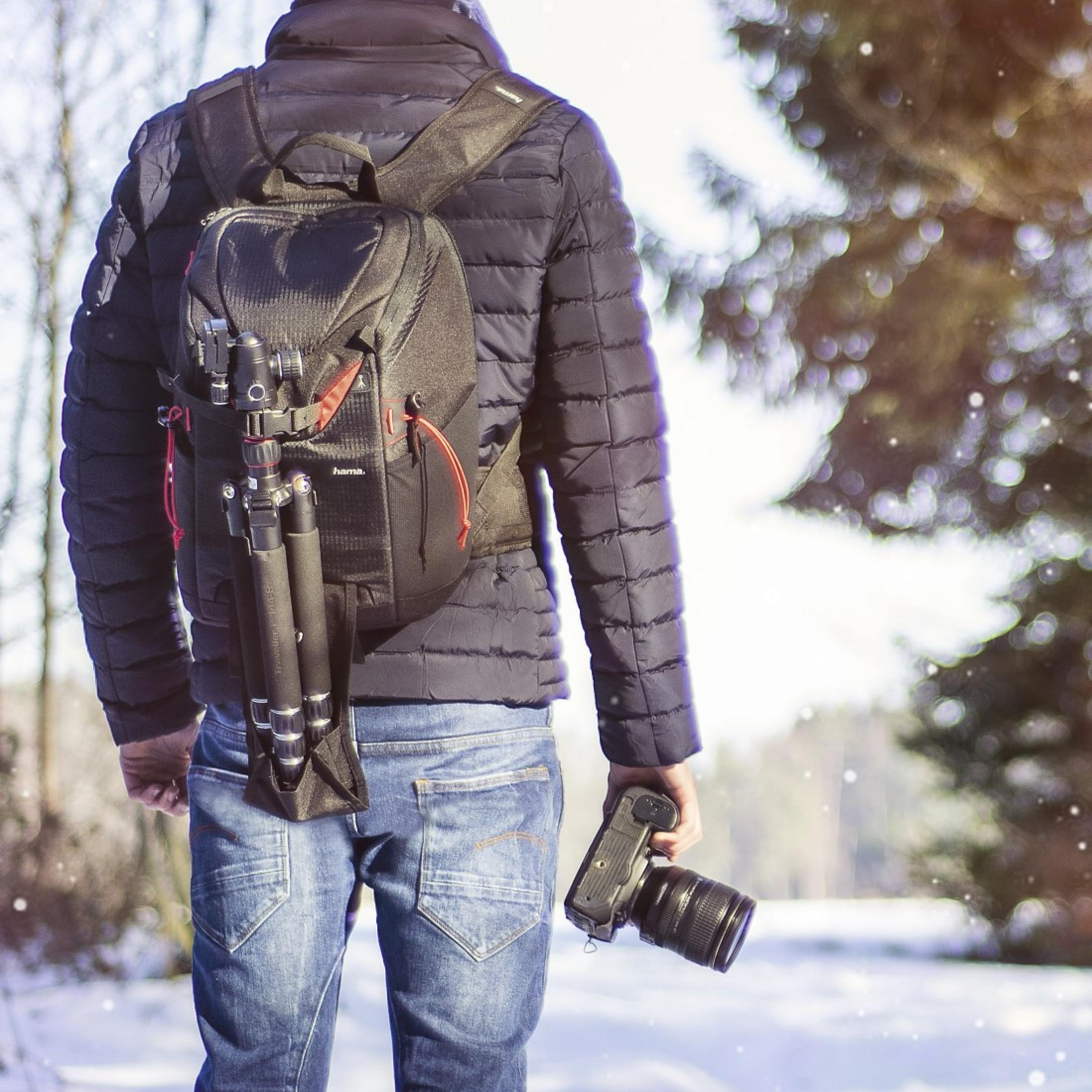 Eine Person mit Rucksack und Kamera mit Stativ, die in einer verschneiten Landschaft läuft.