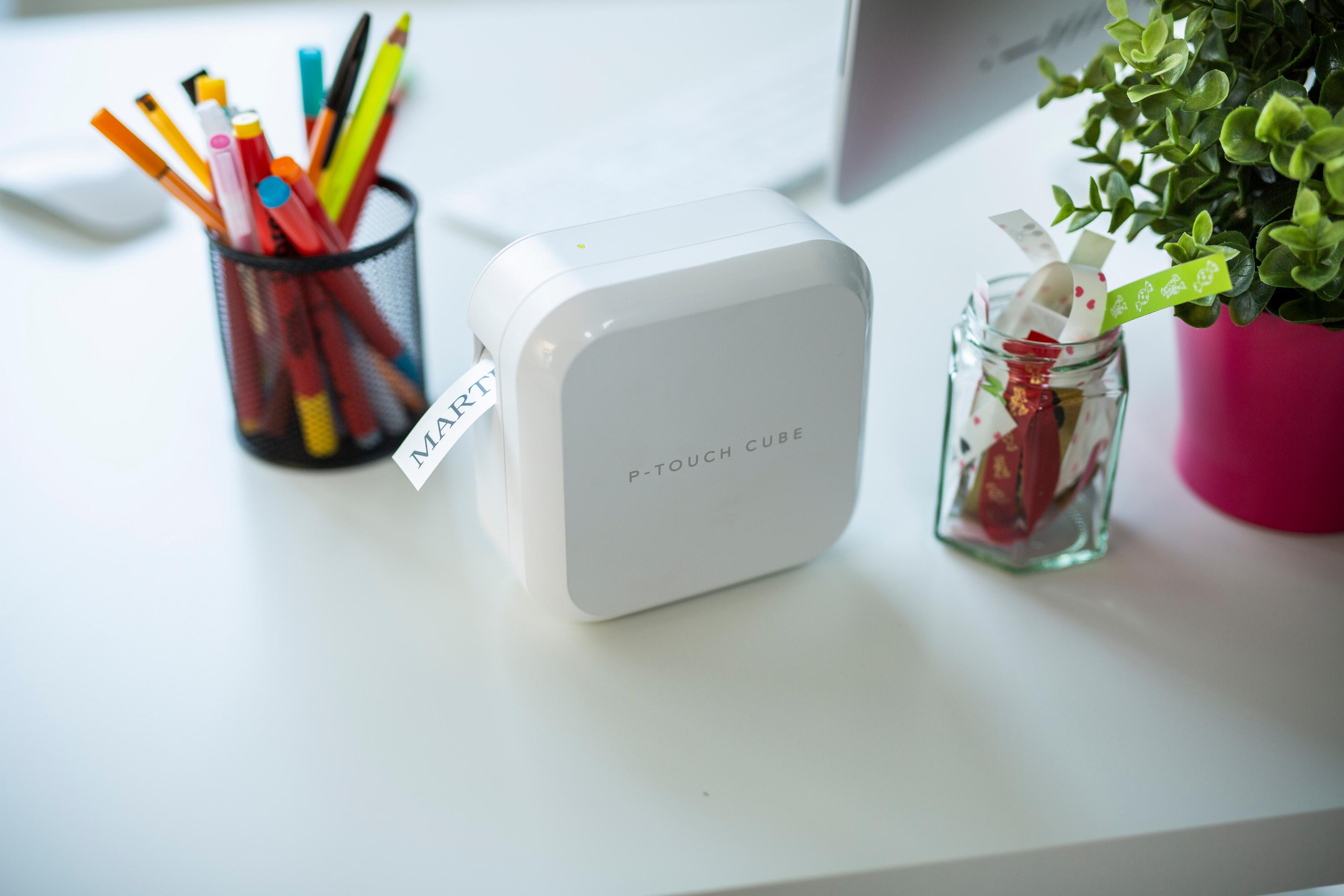 Biały P-Touch Cube na biurku z długopisami, rośliną i oznaczonym słoikiem.