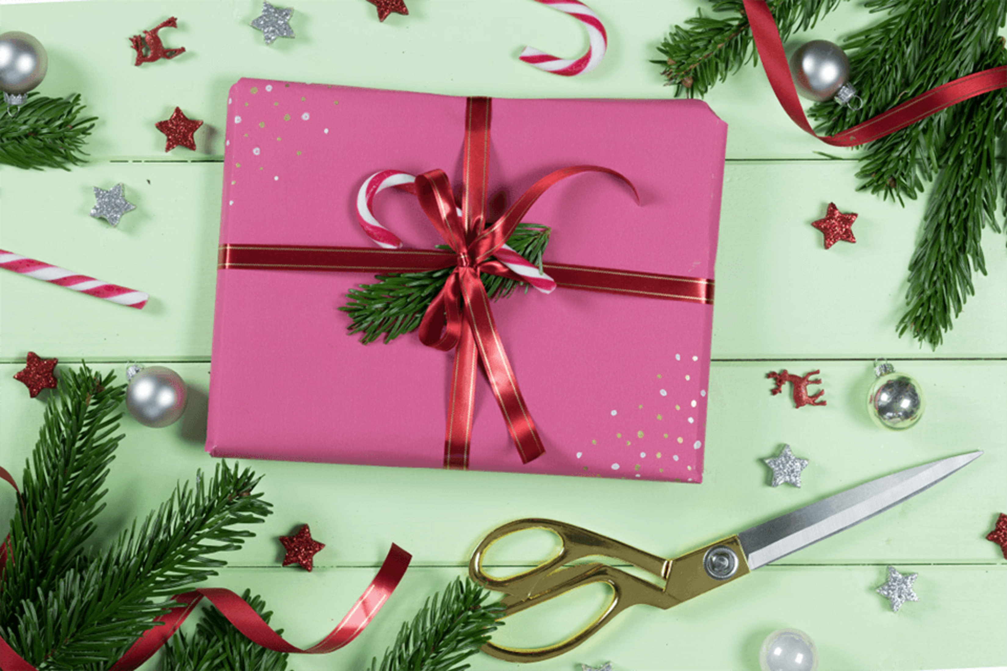 Ein rosa Geschenk, das mit einem roten Band, Zuckerstangen und Grünzeug verpackt ist, mit einer Schere darunter.
