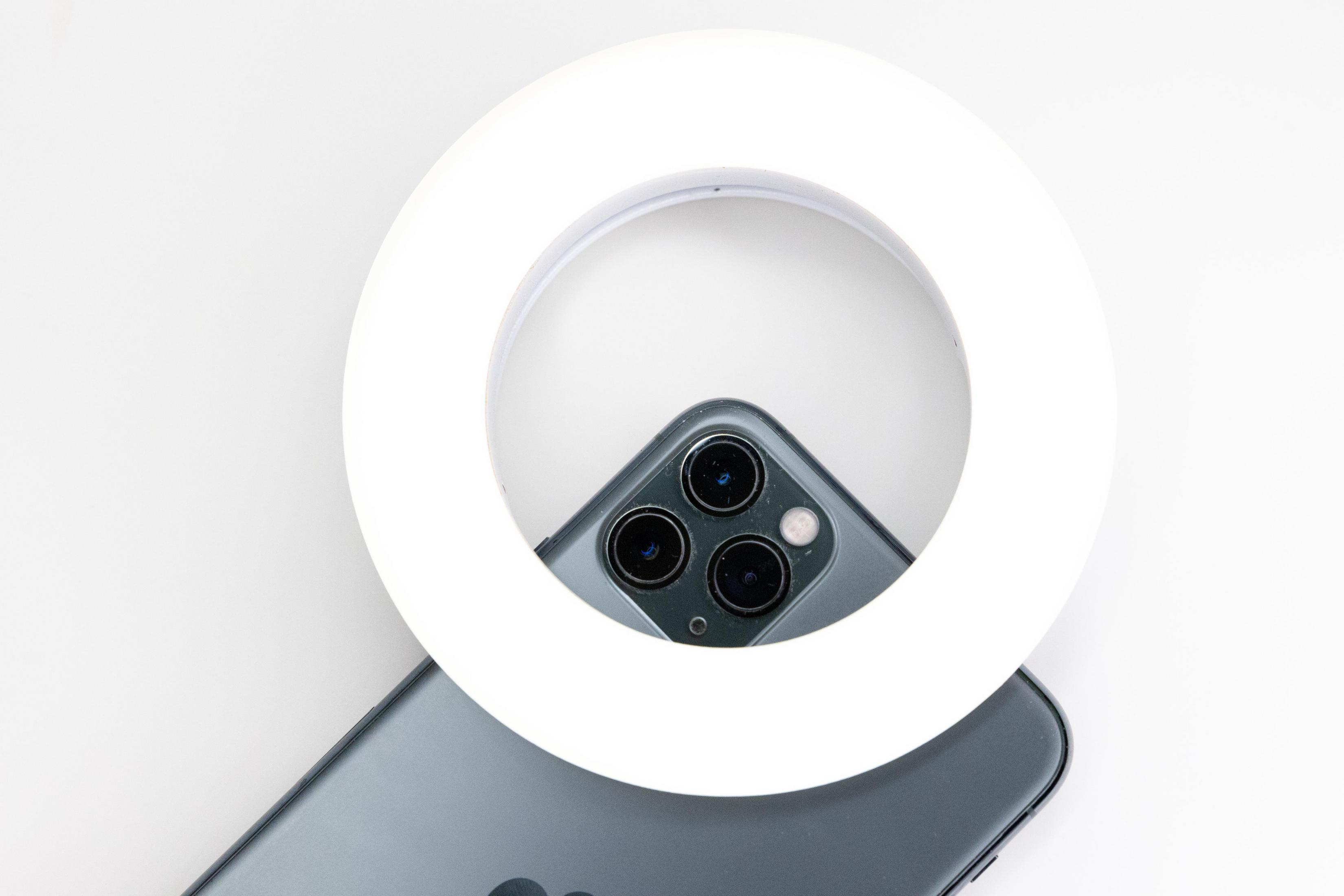 Ein weißes Ringlicht umgibt ein Smartphone mit drei Kameras, auf weißem Hintergrund.