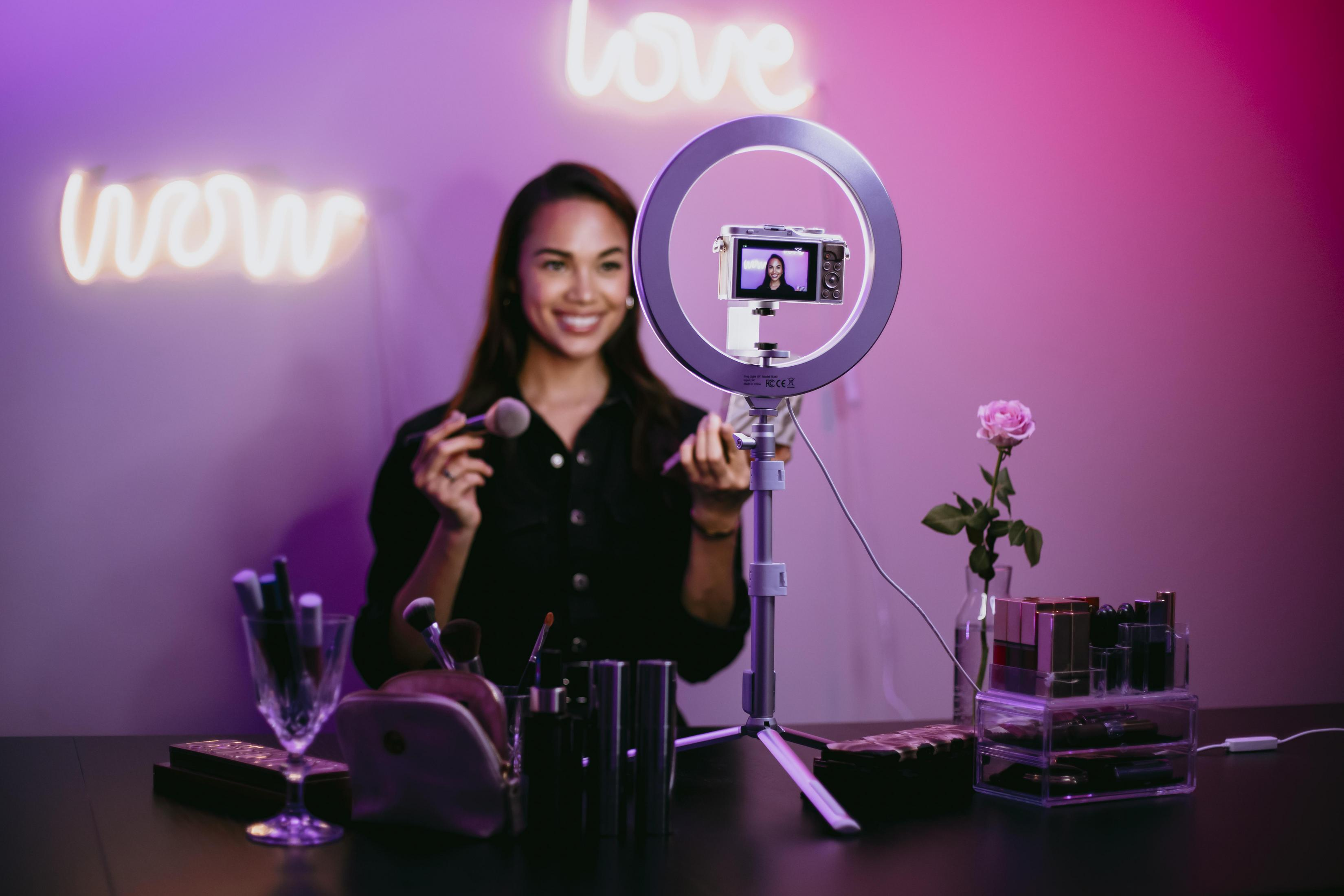 Frau mit Make-up-Pinseln, Ringlicht, Kamera und Neon-Schild, Schönheitsprodukte auf einem Tisch.