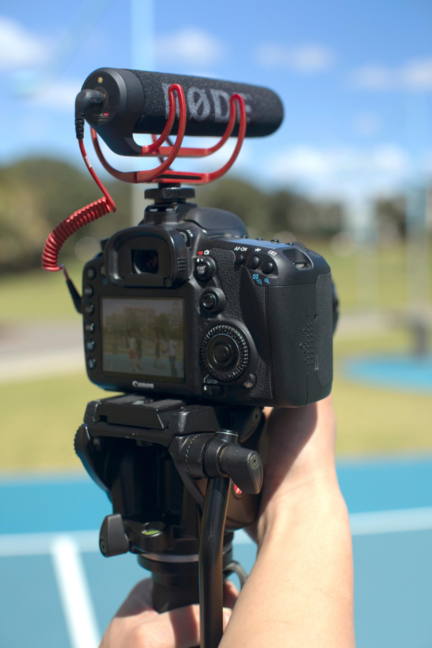 Kamera zamontowana na statywie, z mikrofonem na górze. Tło to błękitne niebo.