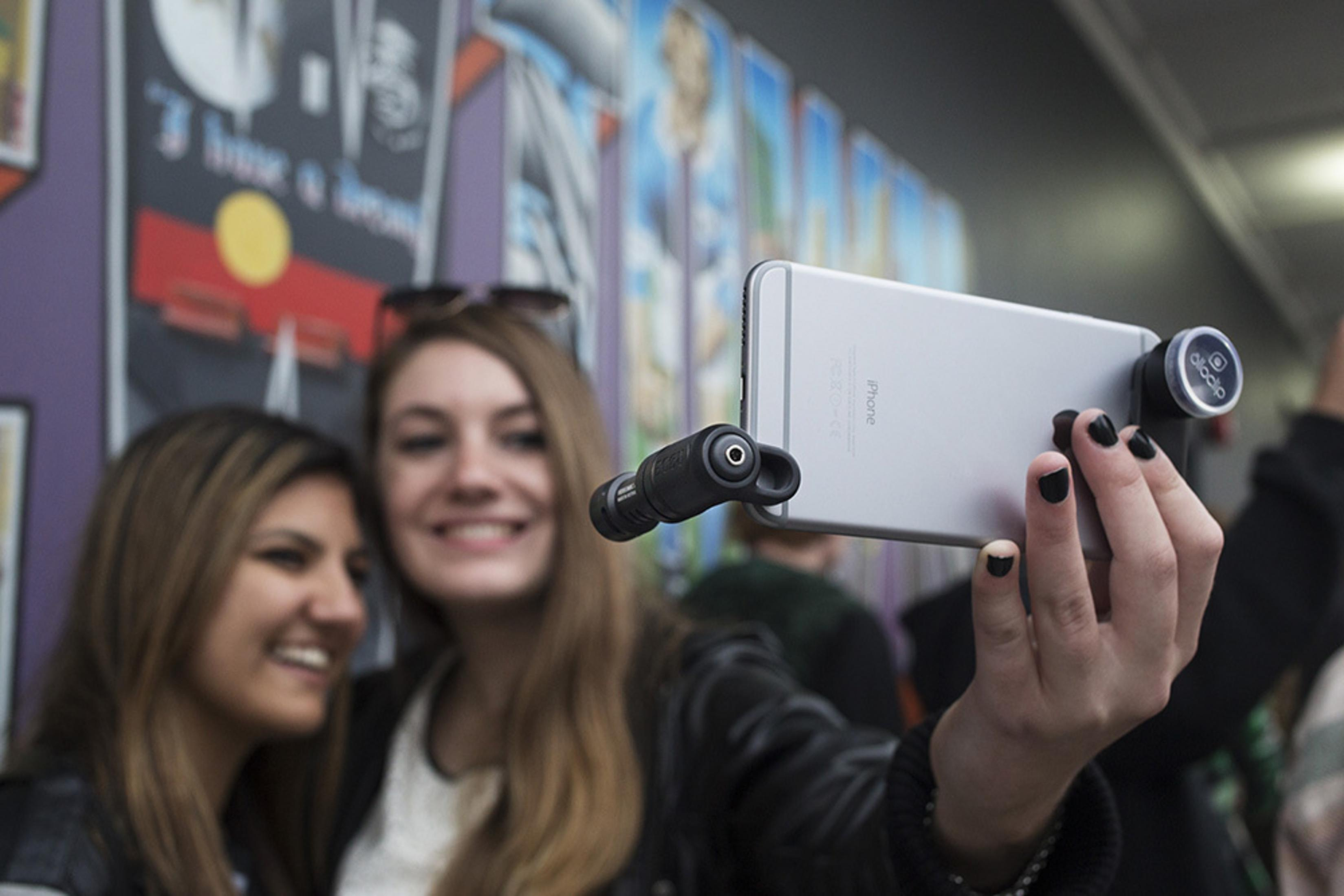 Zwei Frauen machen ein Selfie mit einem Telefon und einem Mikrofon. Sie lächeln. Der Hintergrund hat Graffiti.