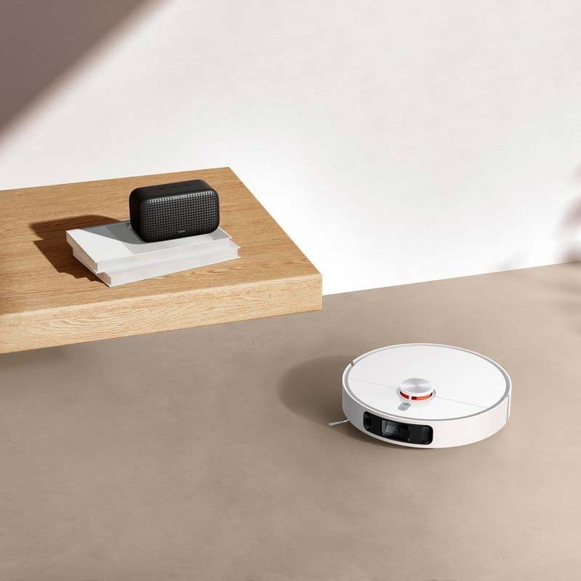 Odkurzacz robot obok małego głośnika na drewnianym stole. Neutralne tło.