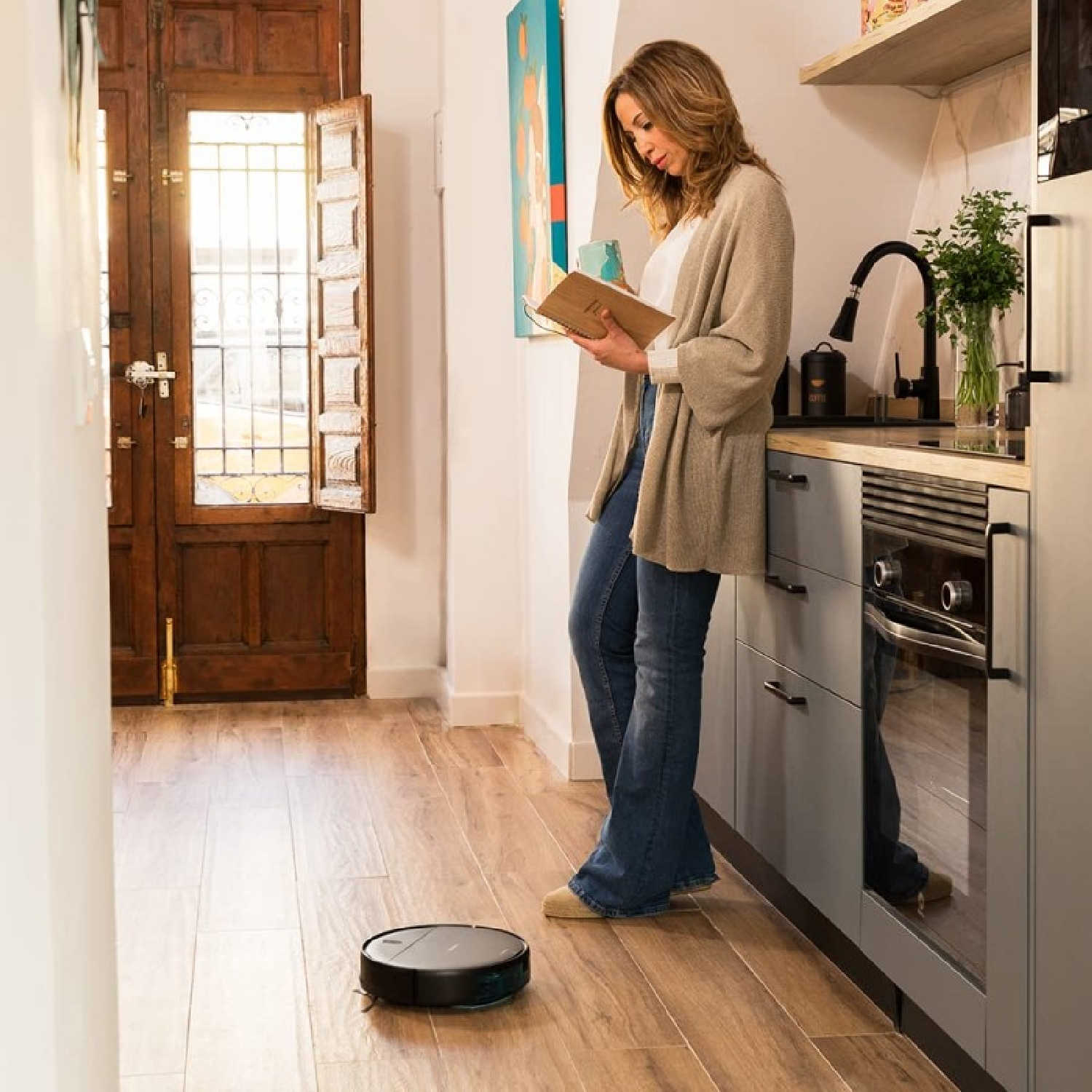 Mujer en la cocina leyendo un libro, una aspiradora robot limpia el suelo.