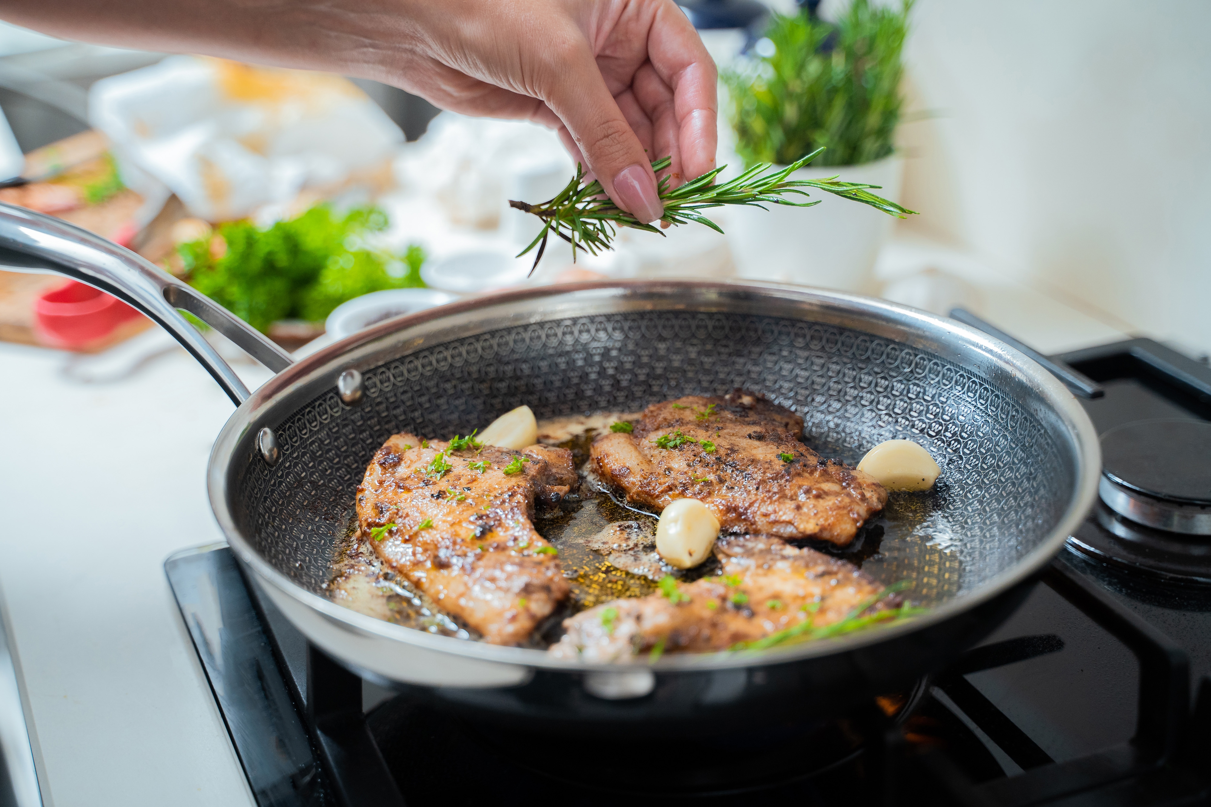 Hand legt Rosmarinzweig auf gebratenes Fleisch in der Pfanne. Knoblauch und Öl sichtbar. Küche.