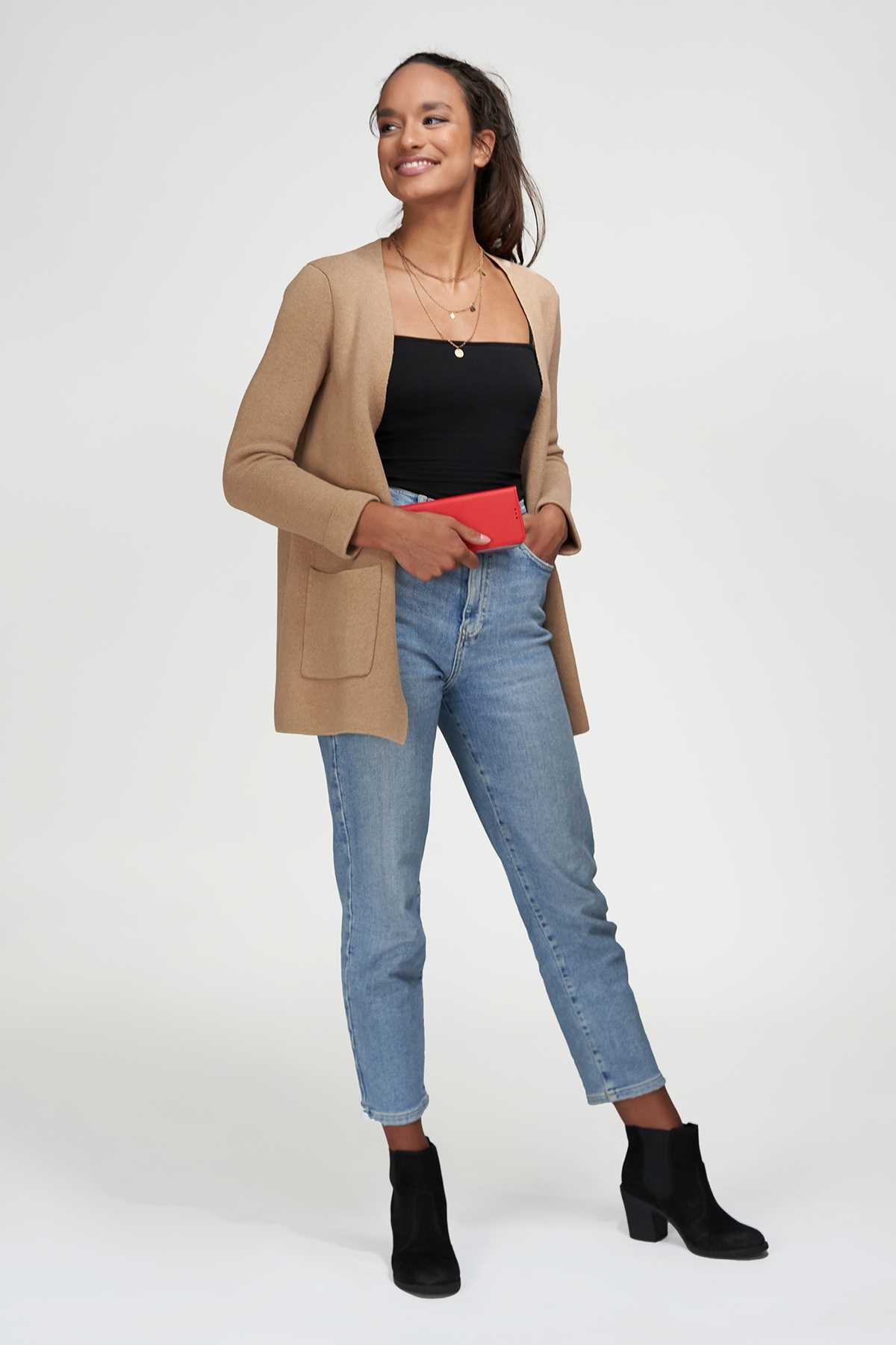 Frau in beigefarbenem Cardigan, schwarzem Top, Jeans, hält rotes Portemonnaie.