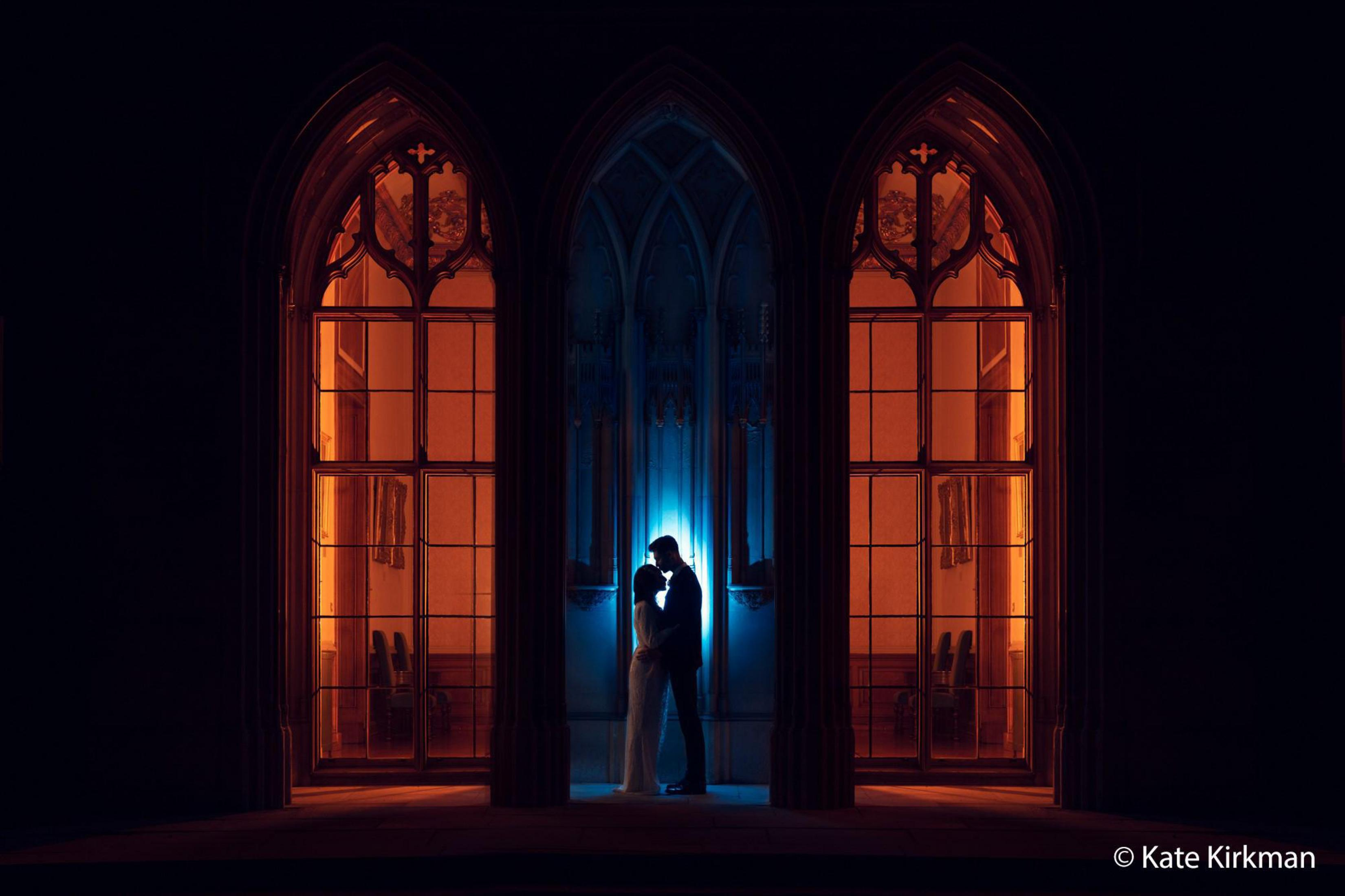 Para w sylwetce przed trzema pomarańczowo oświetlonymi oknami łukowymi.