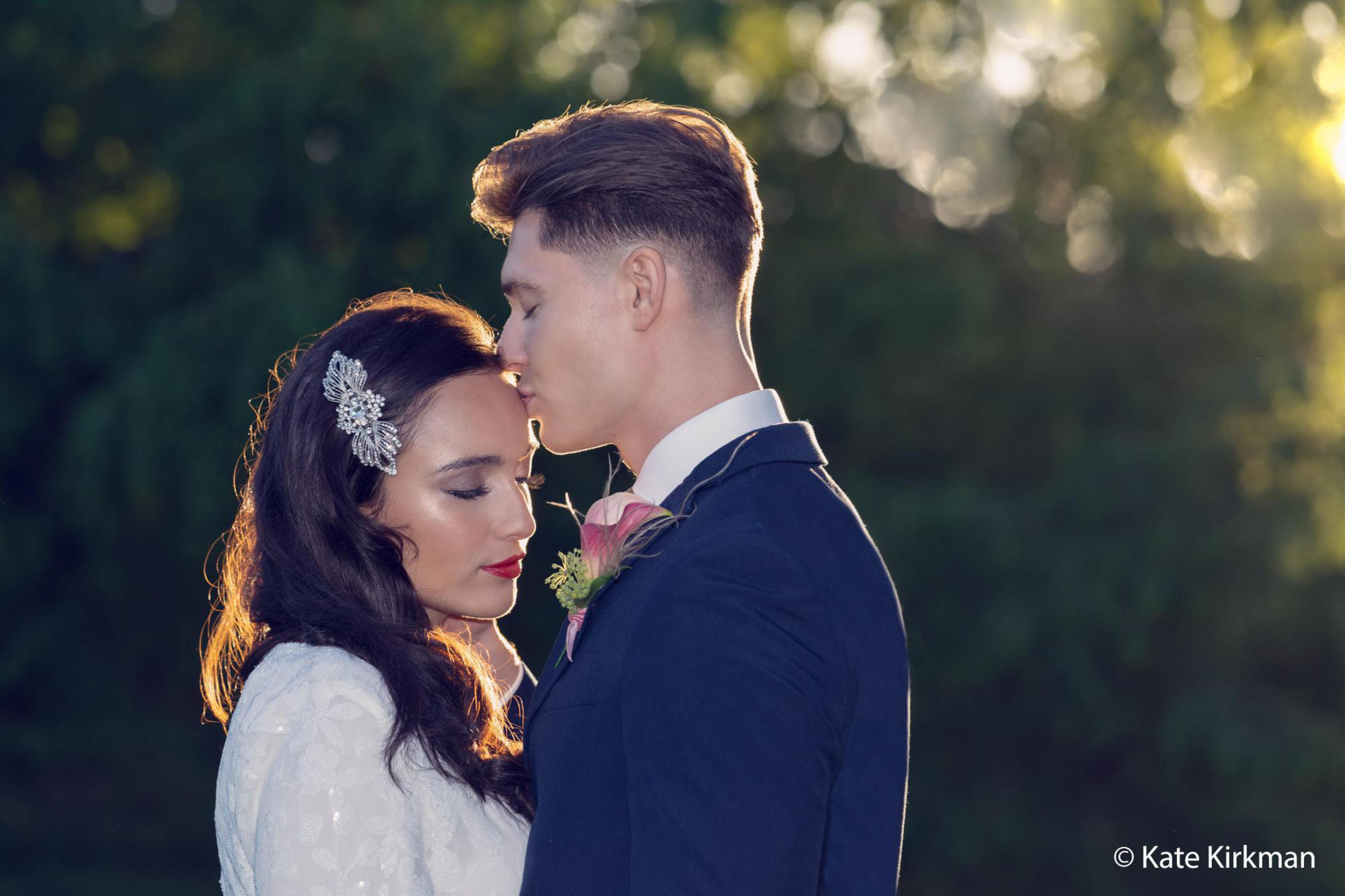 Para obejmująca się. Mężczyzna całuje kobietę w czoło. Kobieta ma ozdobę do włosów i białą suknię.