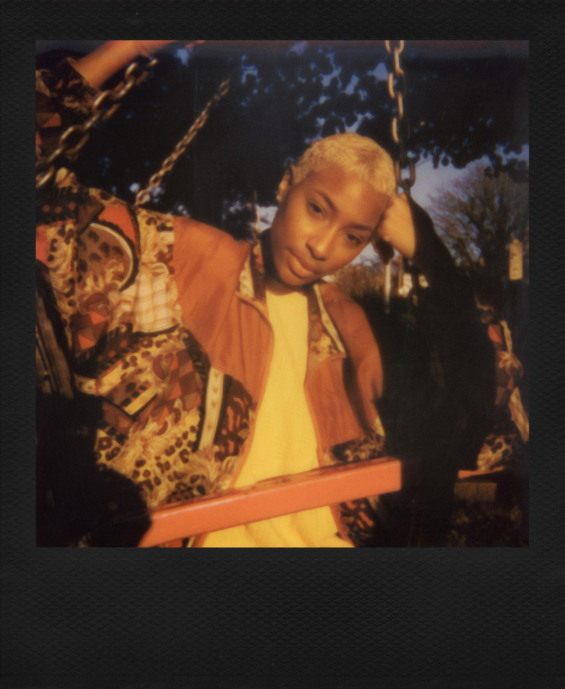 Osoba na huśtawce. Blond włosy, pomarańczowa kurtka i kolorowa koszula. Ciemne tło.