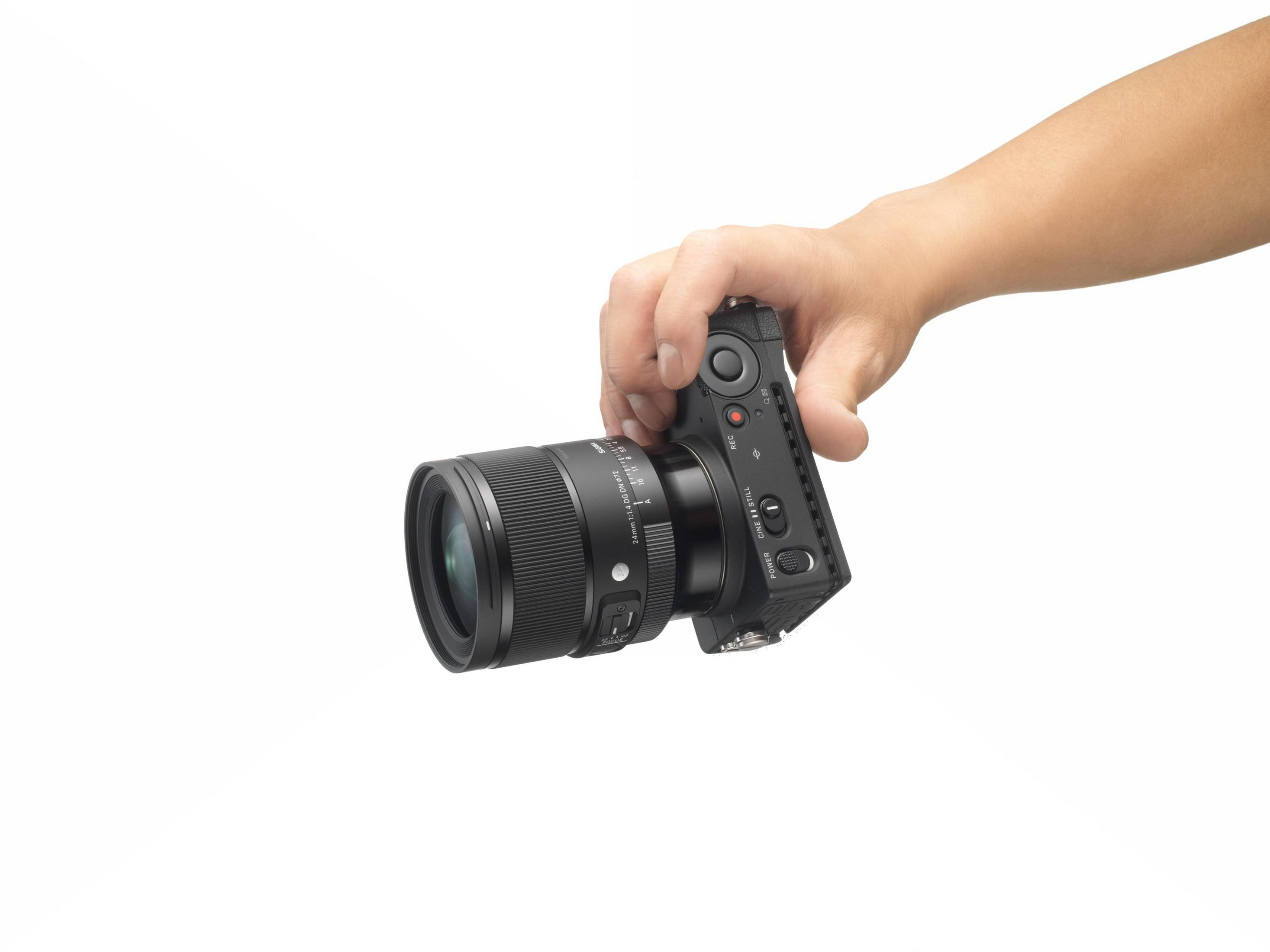Ręka trzymająca aparat z obiektywem, marka Sigma. Białe tło.