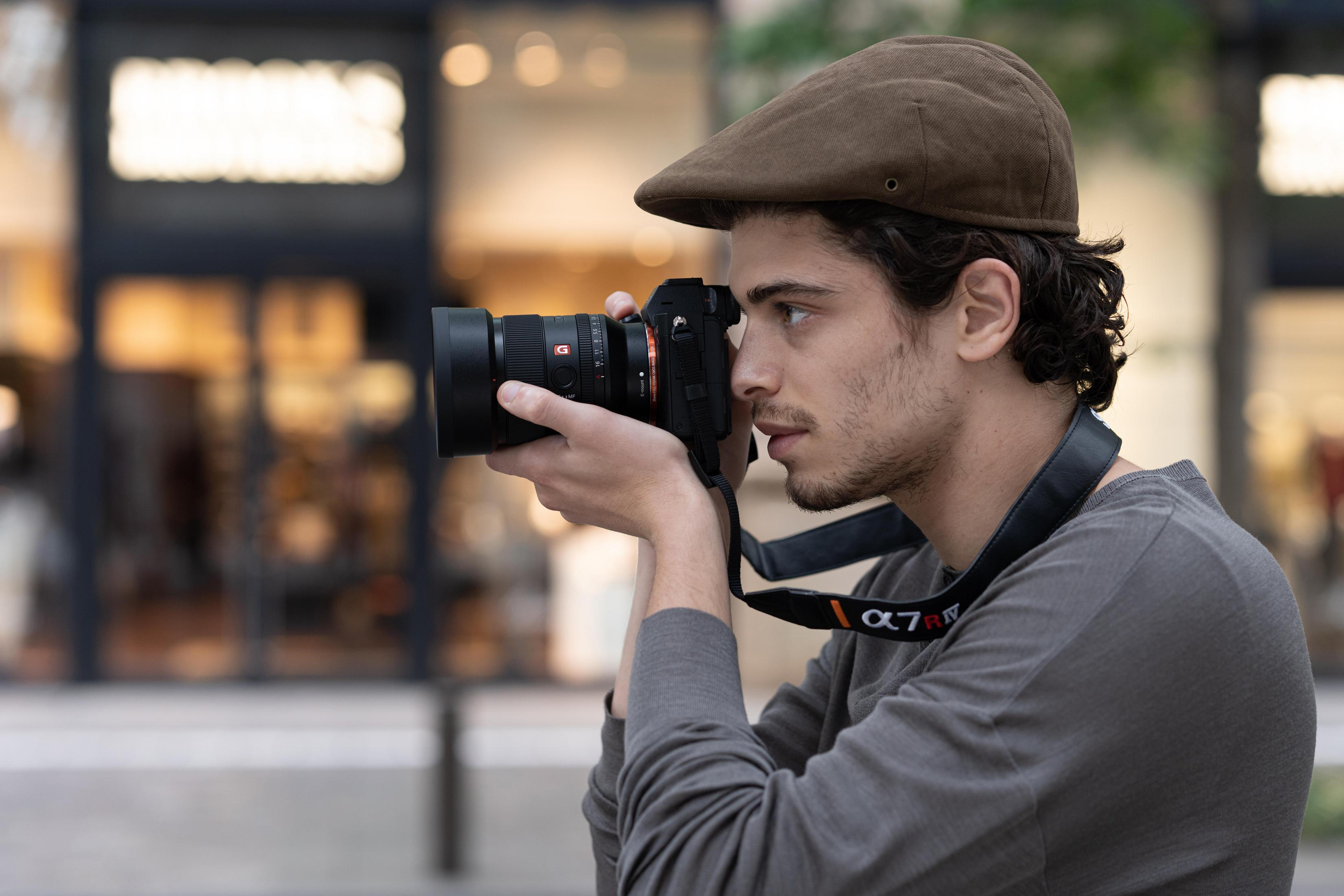 Mężczyzna robiący zdjęcie aparatem, w kapeluszu, na zewnątrz.