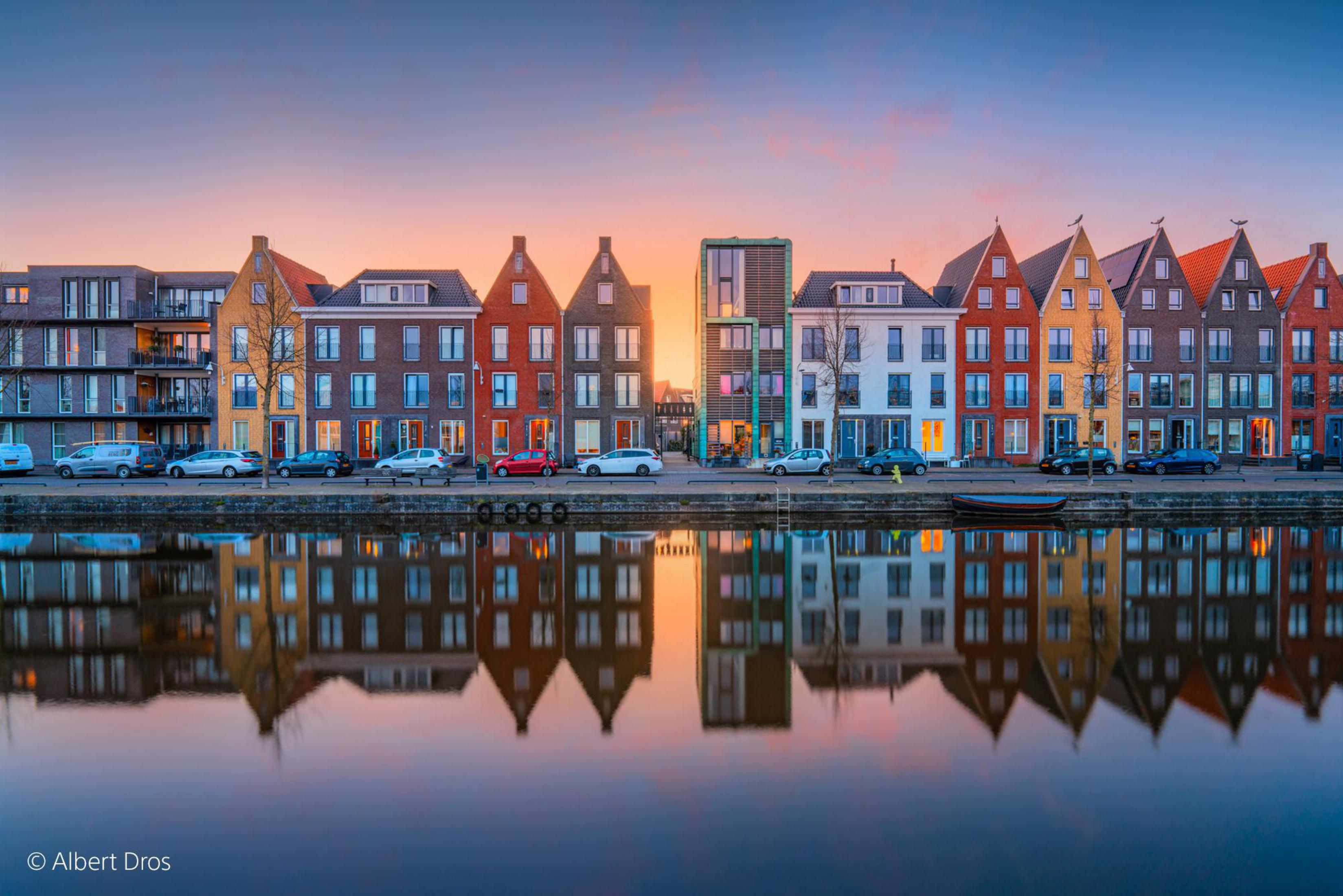 Kolorowe budynki wzdłuż kanału o zachodzie słońca, odbite w wodzie. Samochody zaparkowane.