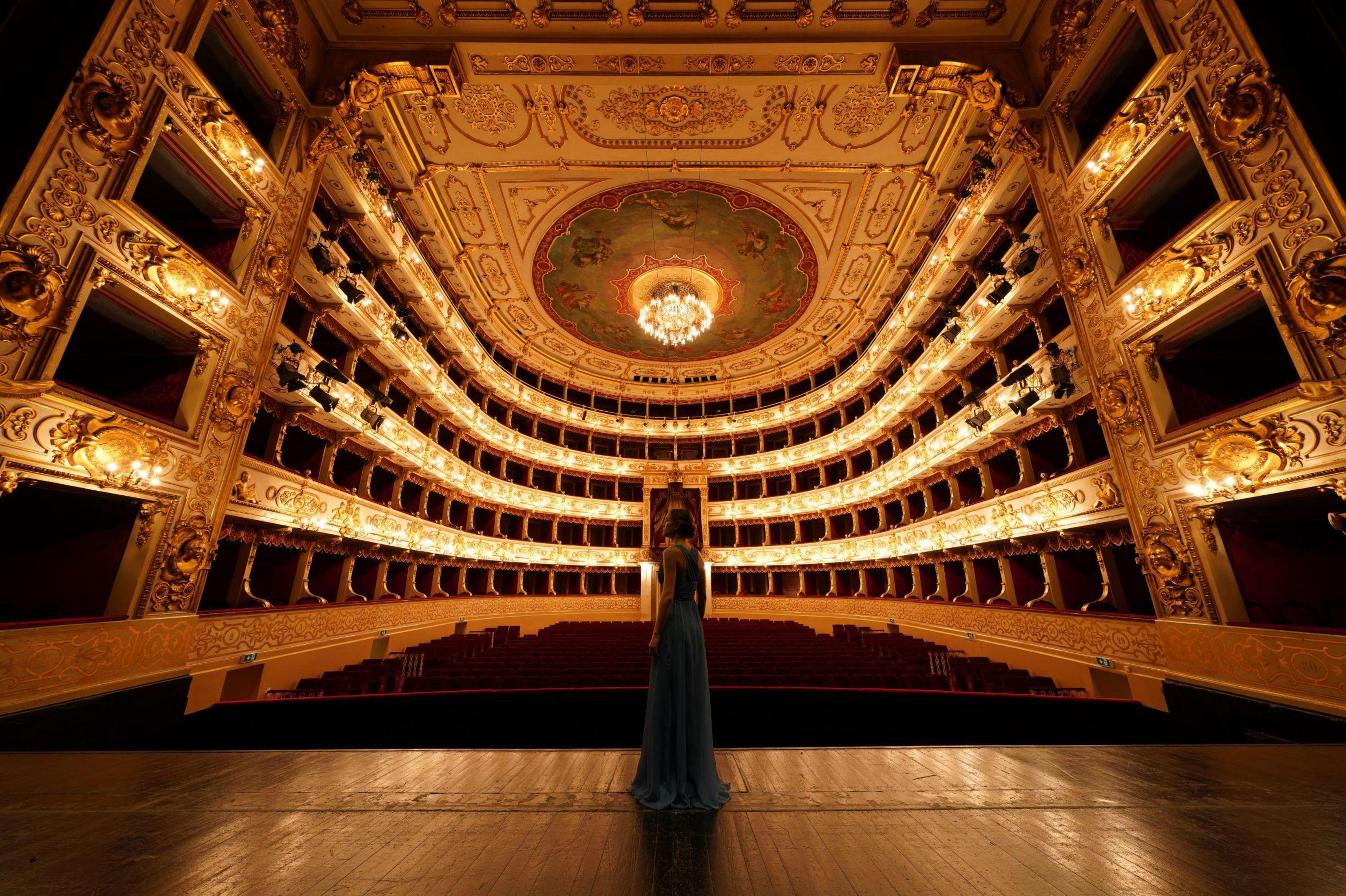Kobieta stoi na scenie w bogato zdobionym teatrze. Fotele są czerwone, sufit złoty.