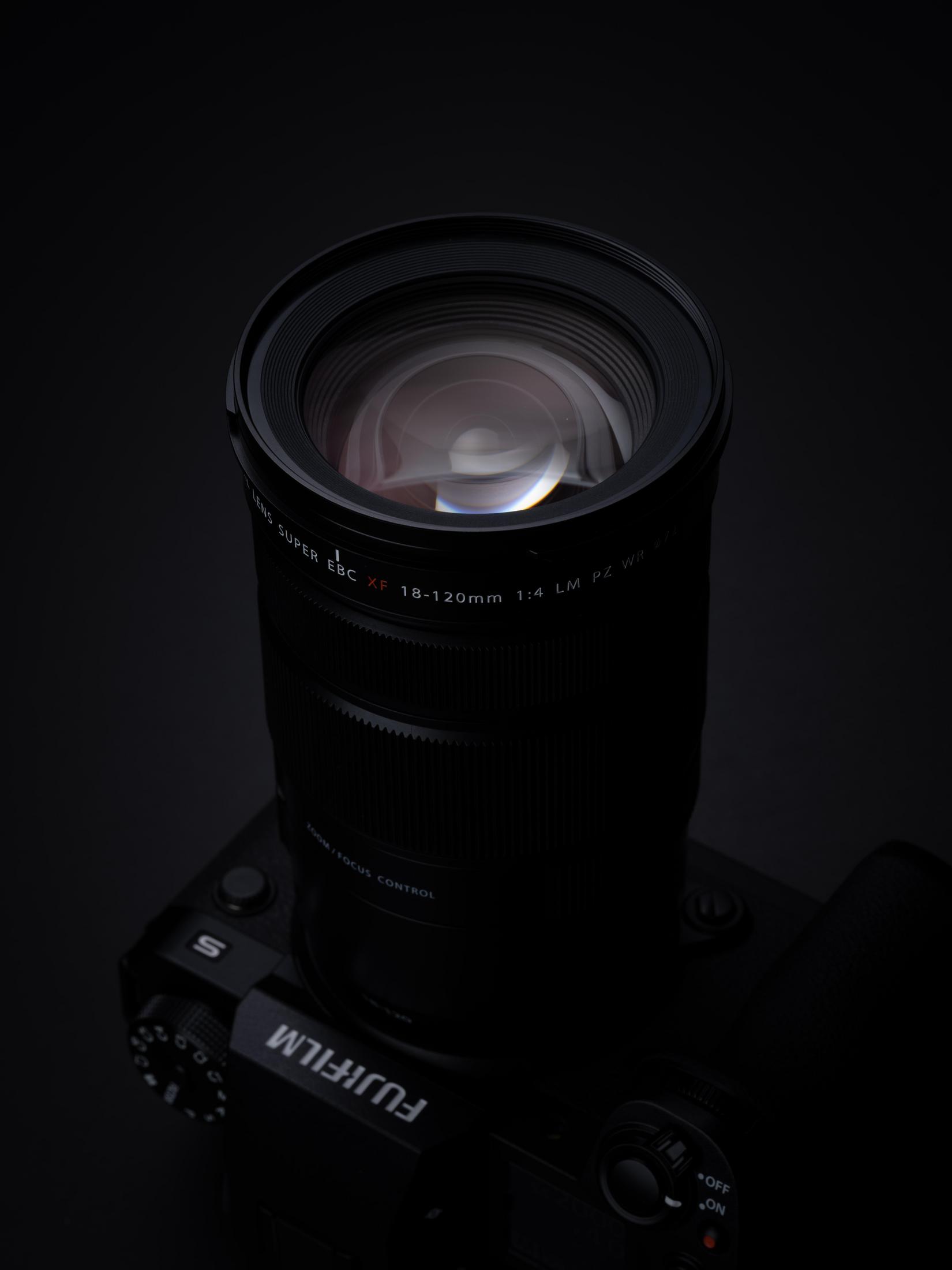 Obiektyw zamontowany na korpusie aparatu. Obiektyw jest czarny z szklanym przodem, ciemne tło.