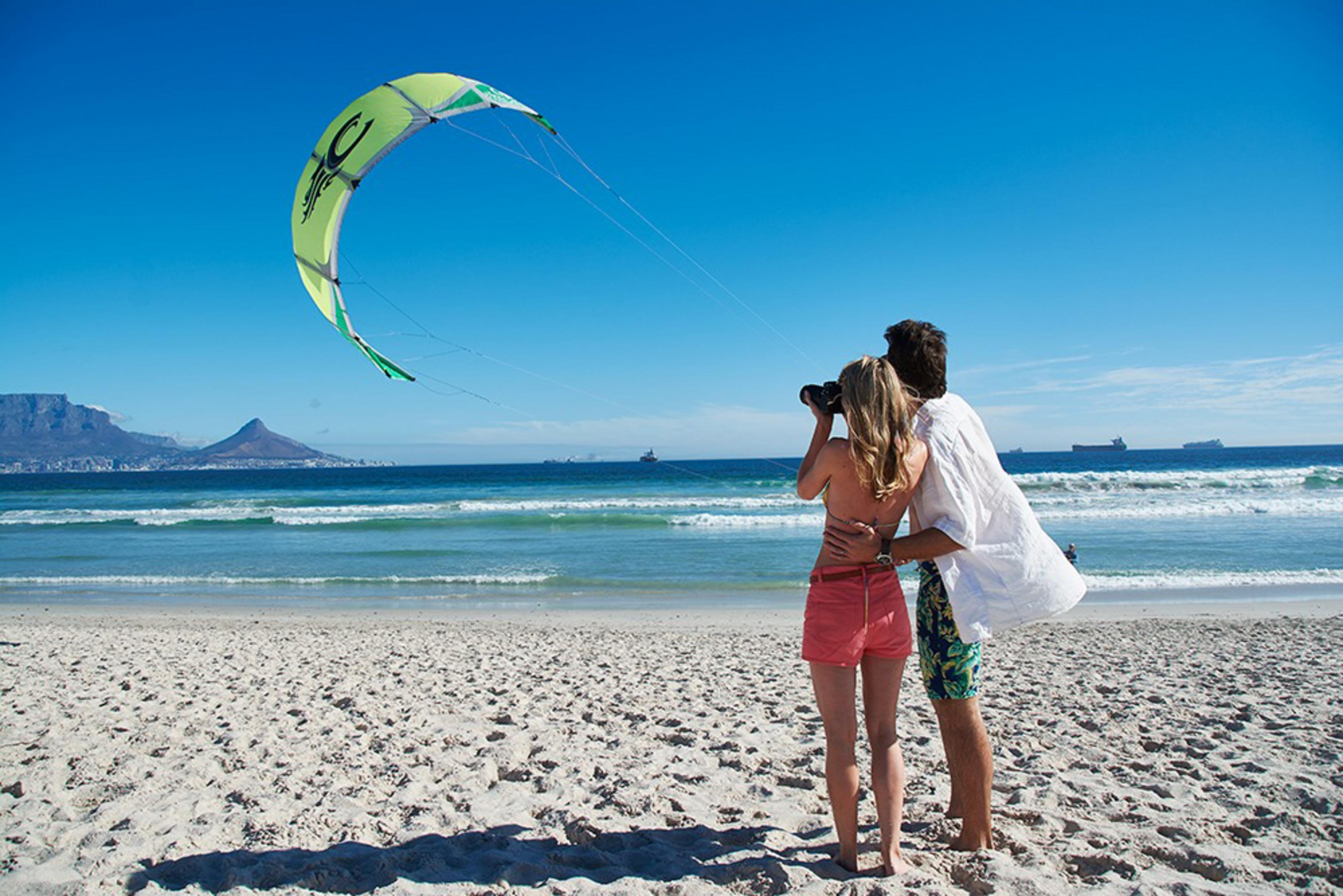 Ein Paar am Strand beobachtet einen Drachen in der Luft. Im Hintergrund ein Berg.