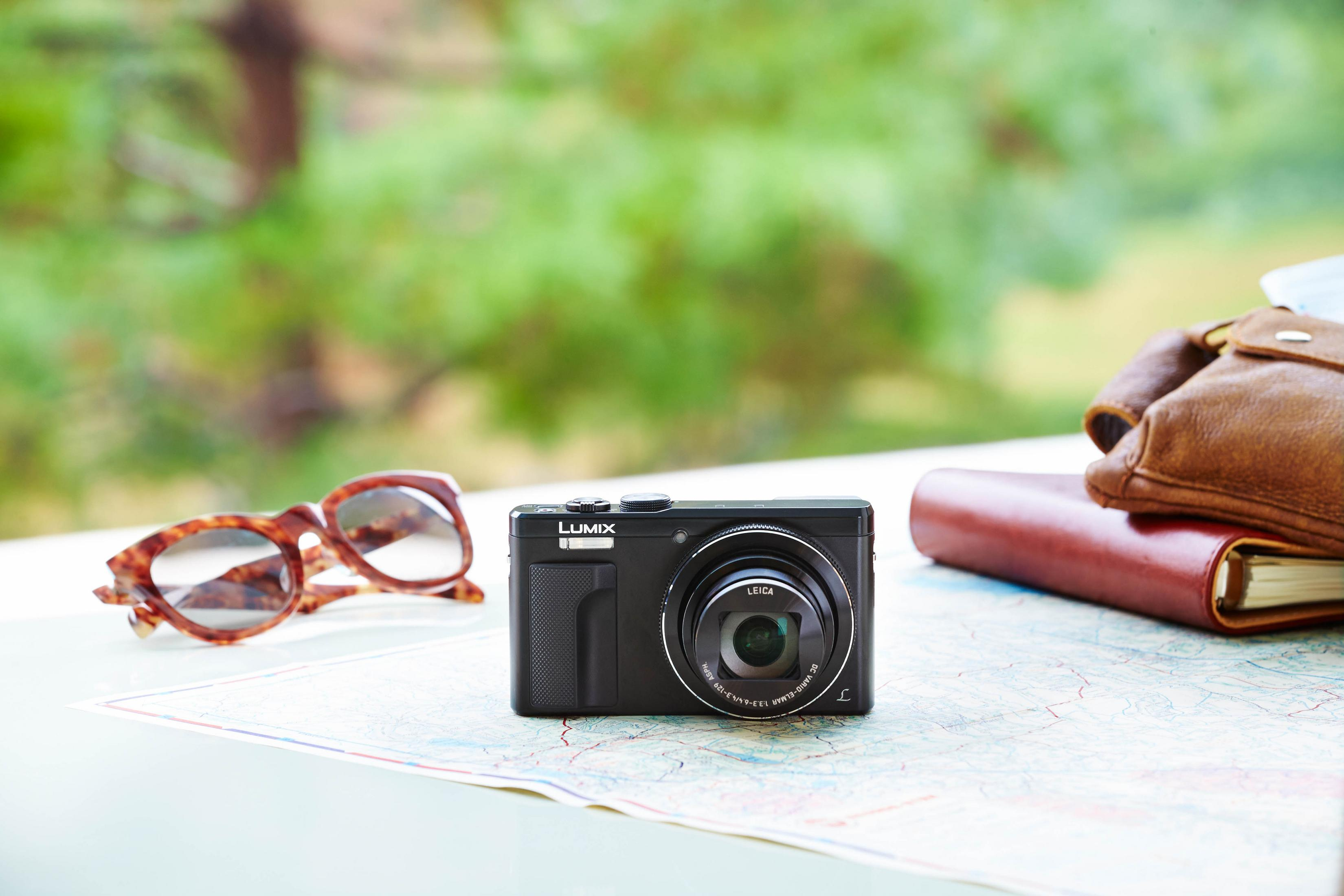 Schwarze Lumix-Kamera auf einer Karte mit Sonnenbrille und Notizbuch, mit grünem Hintergrund.
