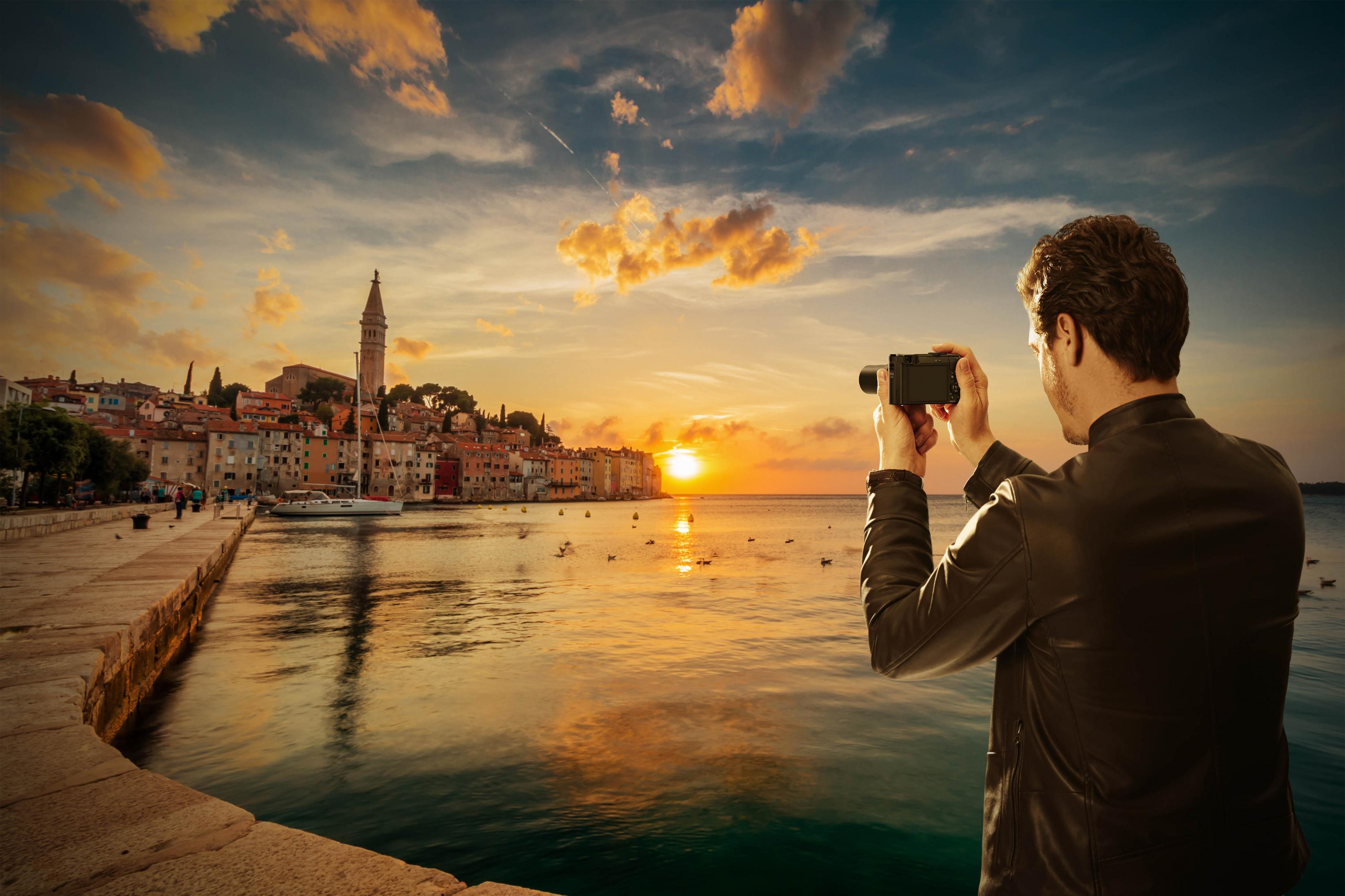 Mann fotografiert Küstenstadt bei Sonnenuntergang. Gebäude, Wasser und Himmel sind sichtbar.