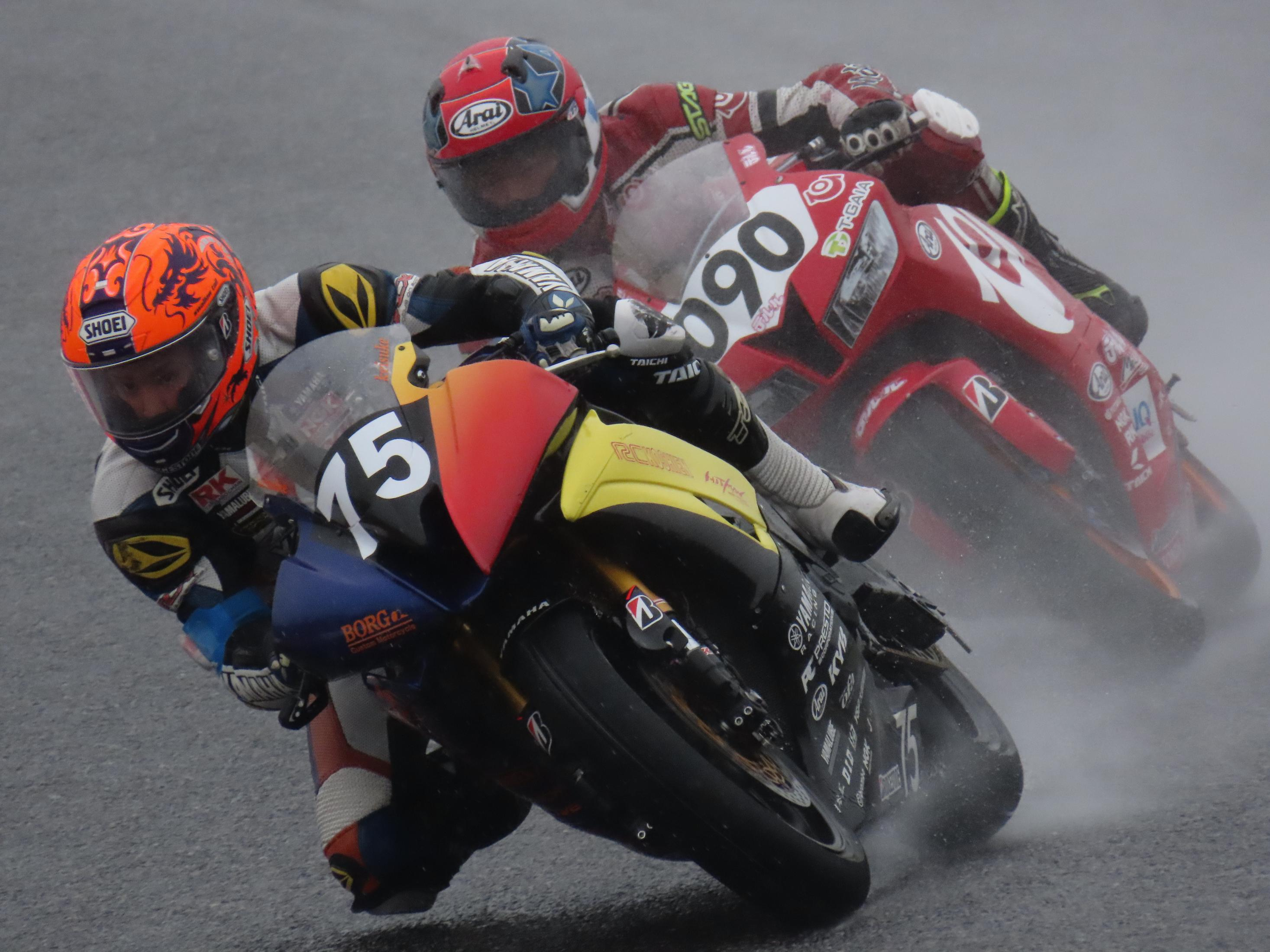 Piloti di moto su pista bagnata, primo piano, con sfondo sfocato durante una gara piovosa.