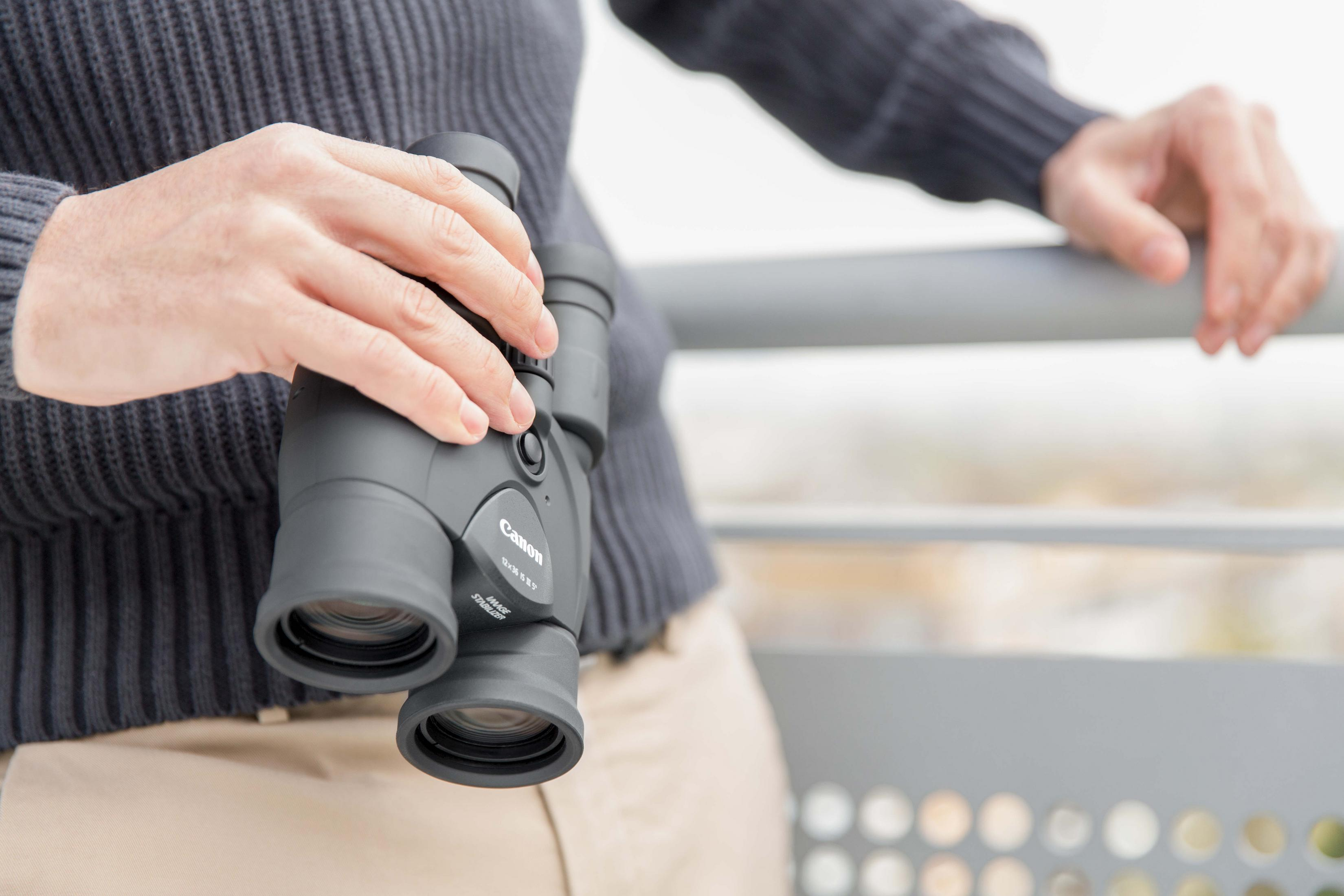 Osoba trzymająca czarną lornetkę. Ma na sobie niebieski sweter i beżowe spodnie.
