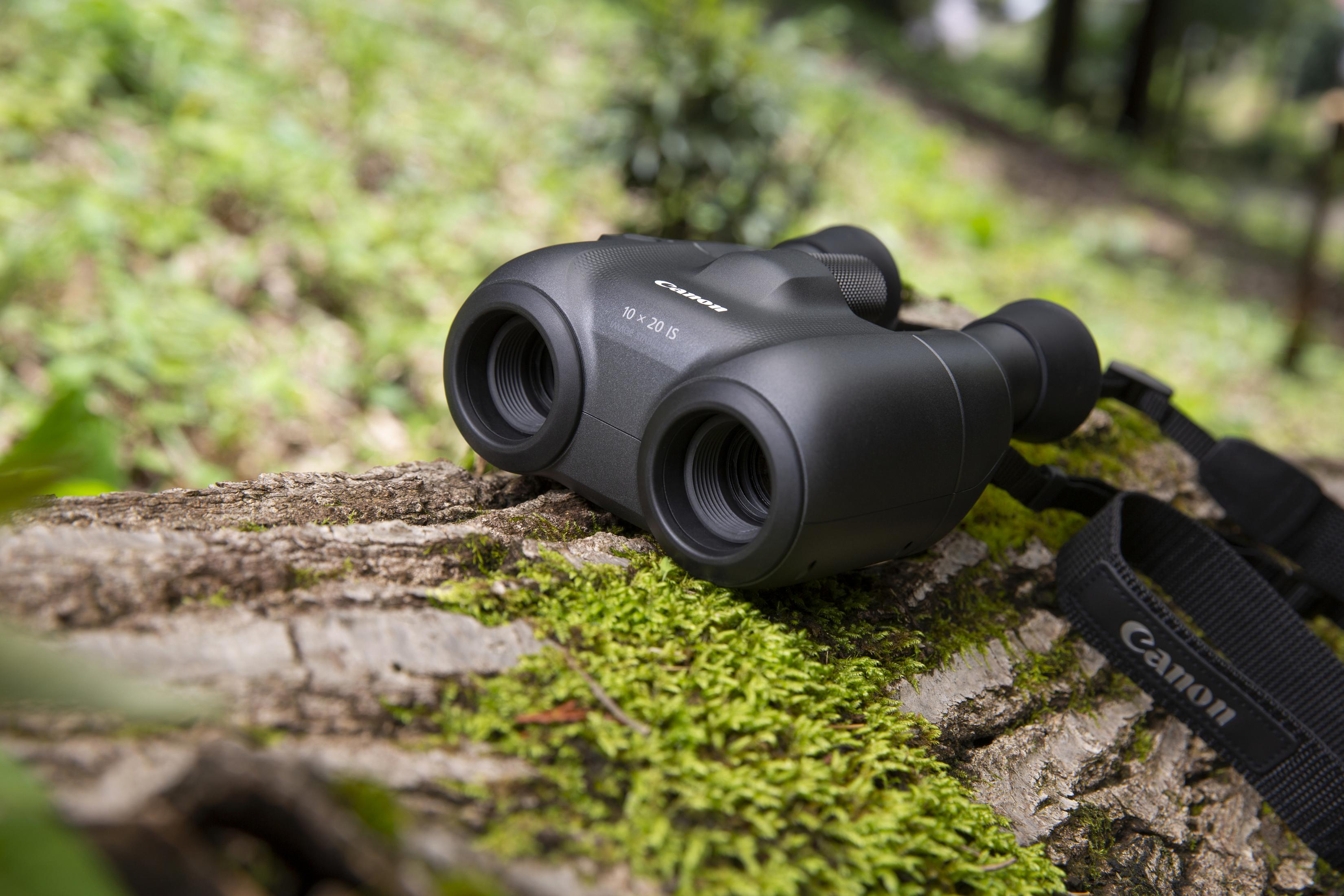 Czarna lornetka Canon, spoczywająca na pniu drzewa pokrytym zielonym mchem. Widoczny jest czarny pasek.