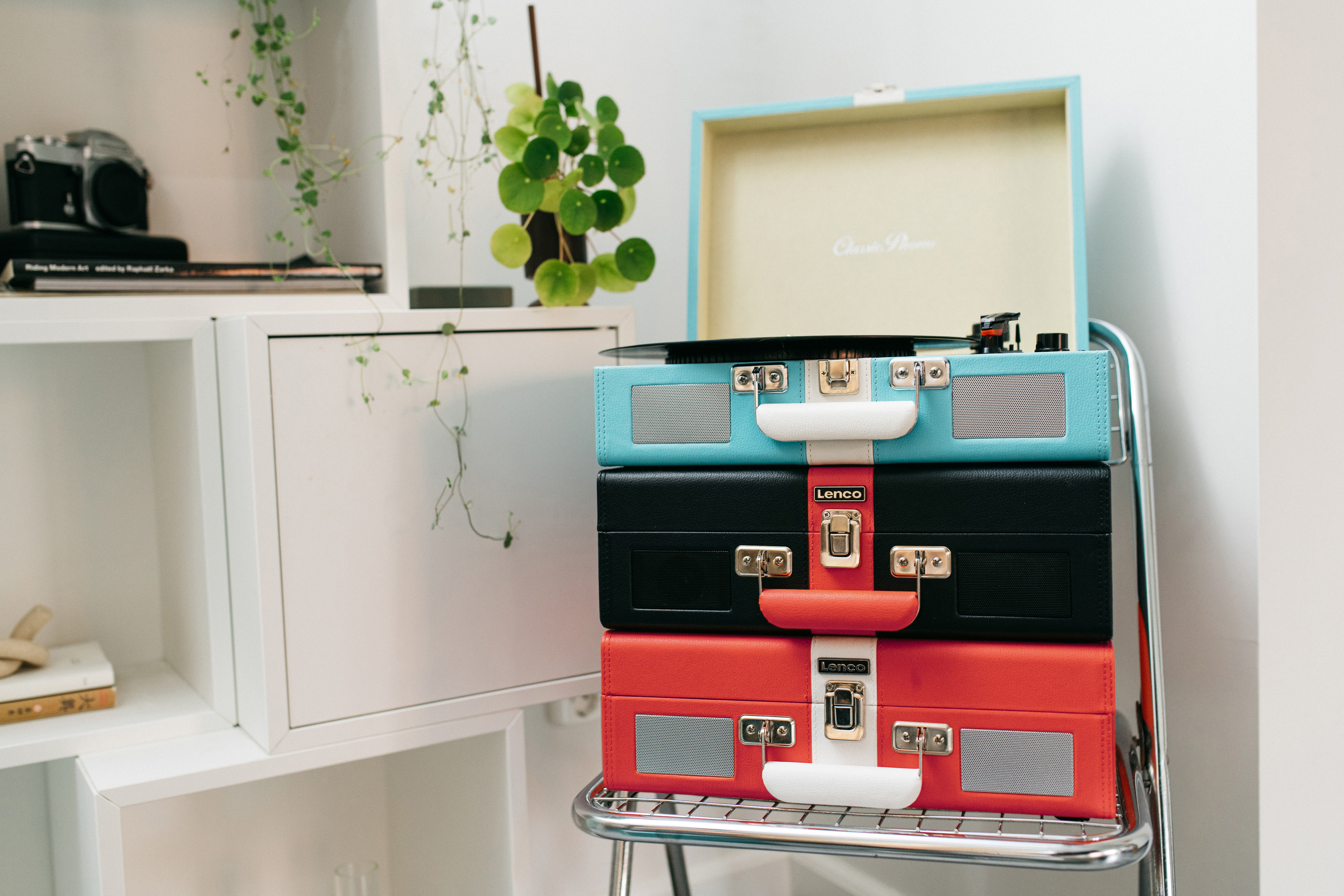 Trzy ułożone w stosy gramofony w stylu vintage w kolorze niebieskim, czarnym i czerwonym. W tle półka z roślinami i biała szafka.