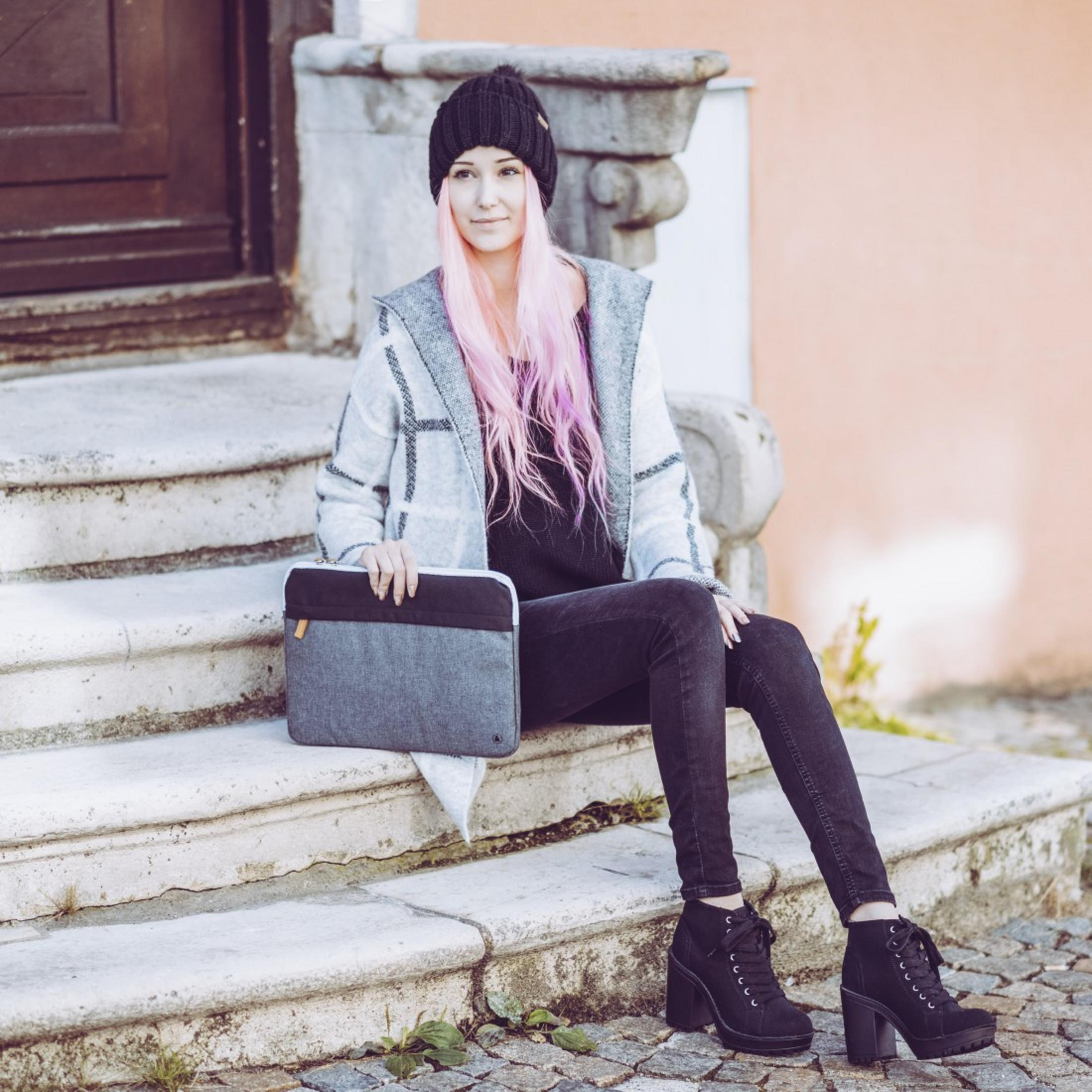 Kobieta z różowymi włosami siedzi na schodach, trzymając szarą torbę na laptopa. Ma na sobie czarną czapkę i czarne buty.