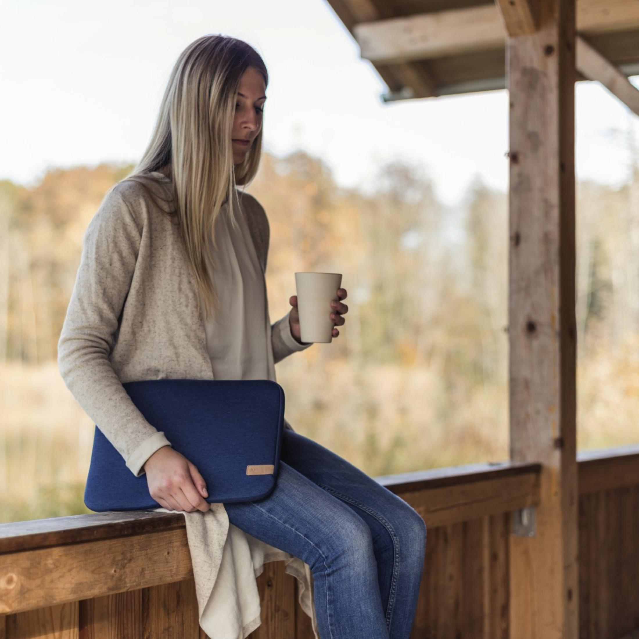 Kobieta siedzi, trzyma kubek, z laptopem w niebieskim etui. Scena na drewnianym tarasie.