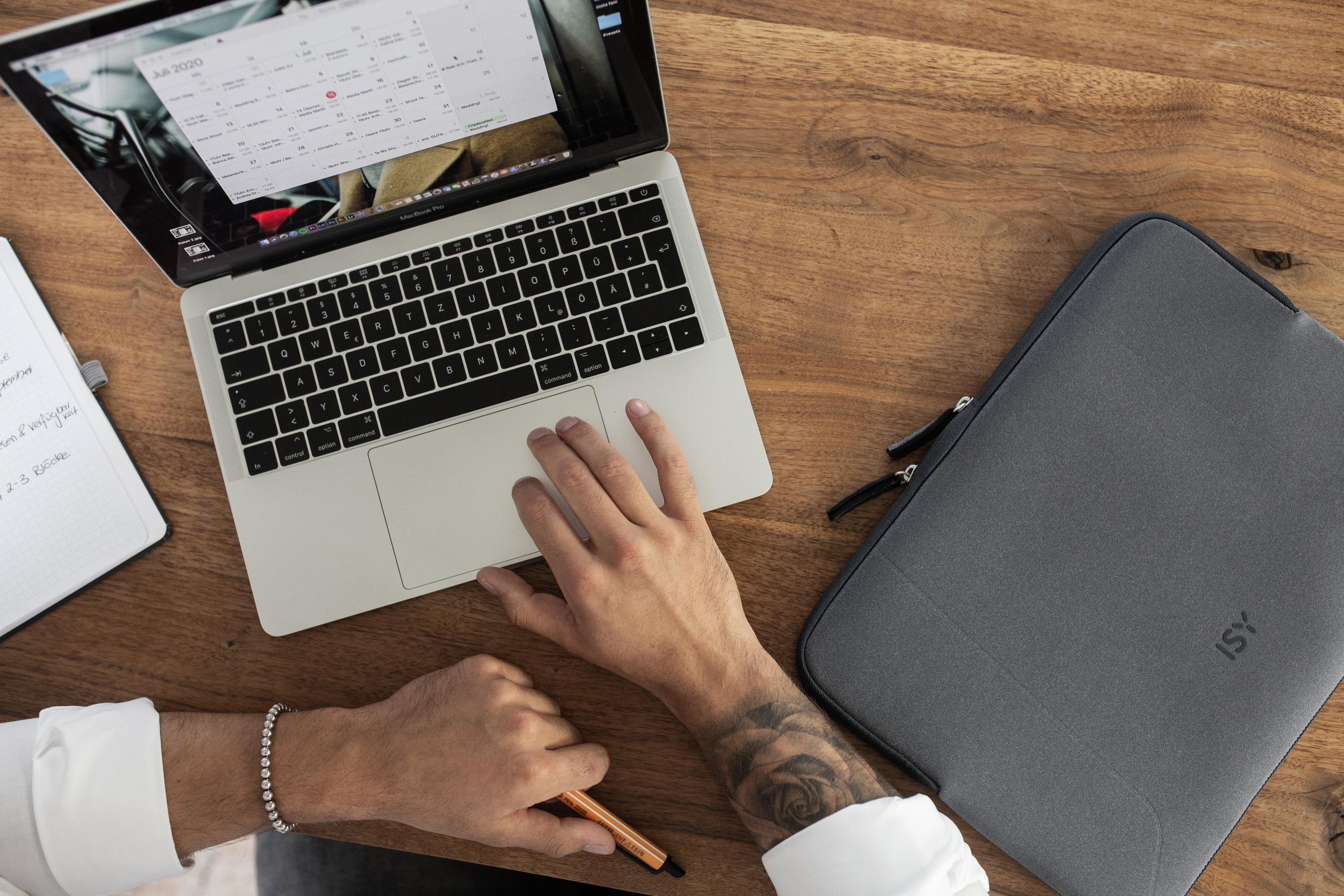 Eine Person benutzt ein MacBook auf einem Holztisch. Daneben liegen ein Notizbuch, Stift und eine Laptop-Tasche.
